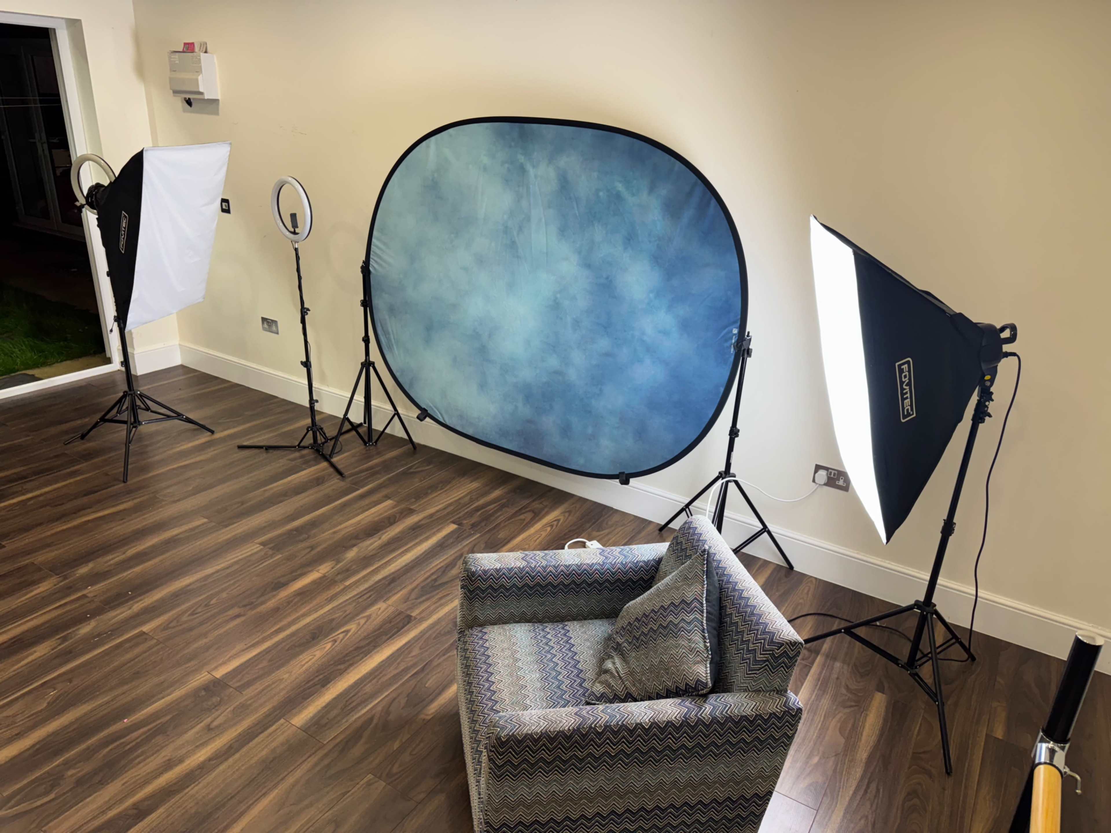 The image shows a sparsely furnished studio space with a blue backdrop, lighting equipment, and an armchair arranged on a wooden floor.