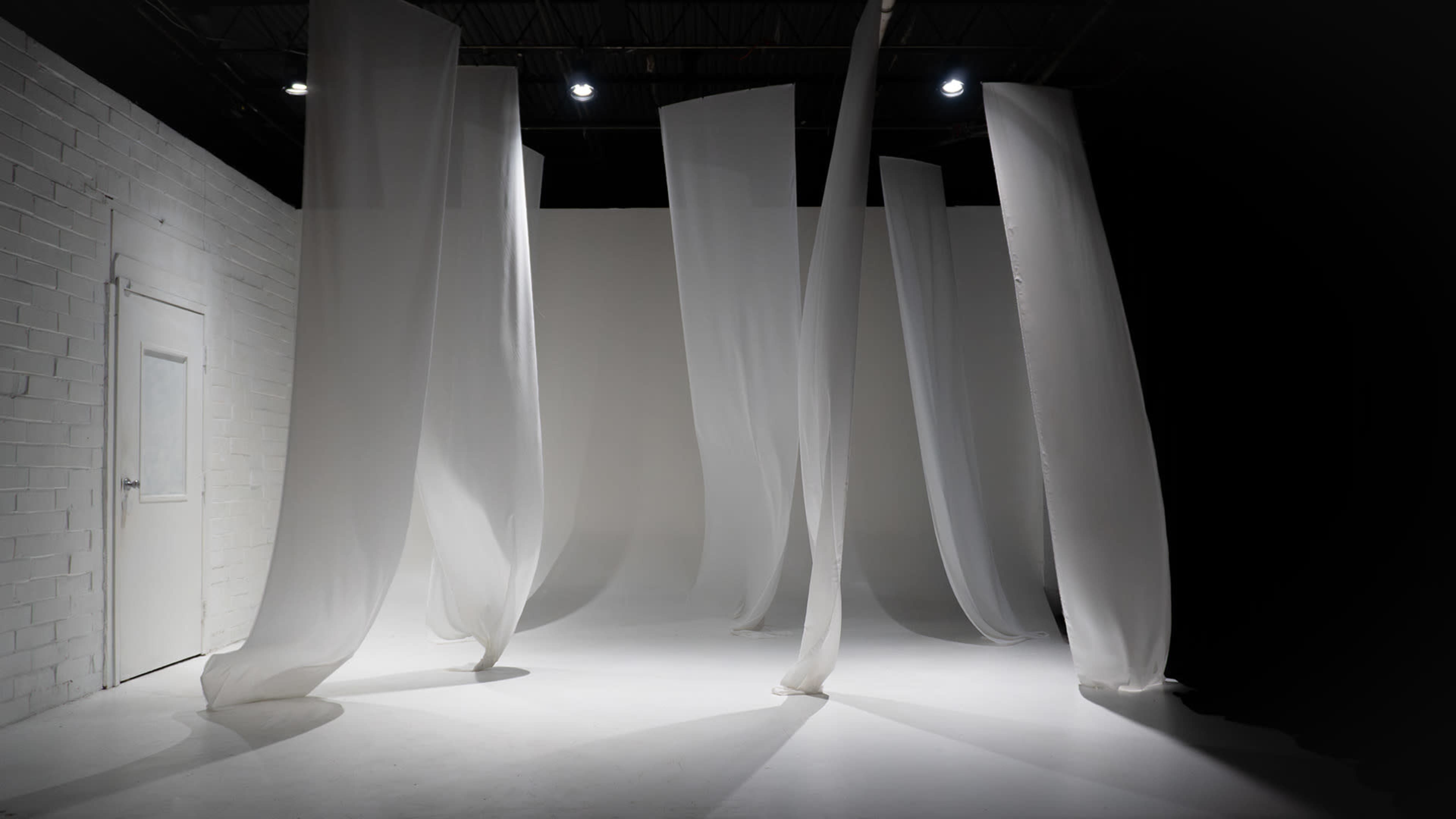 The image shows a minimalistic studio space with flowing white curtains suspended from the ceiling against a stark backdrop.