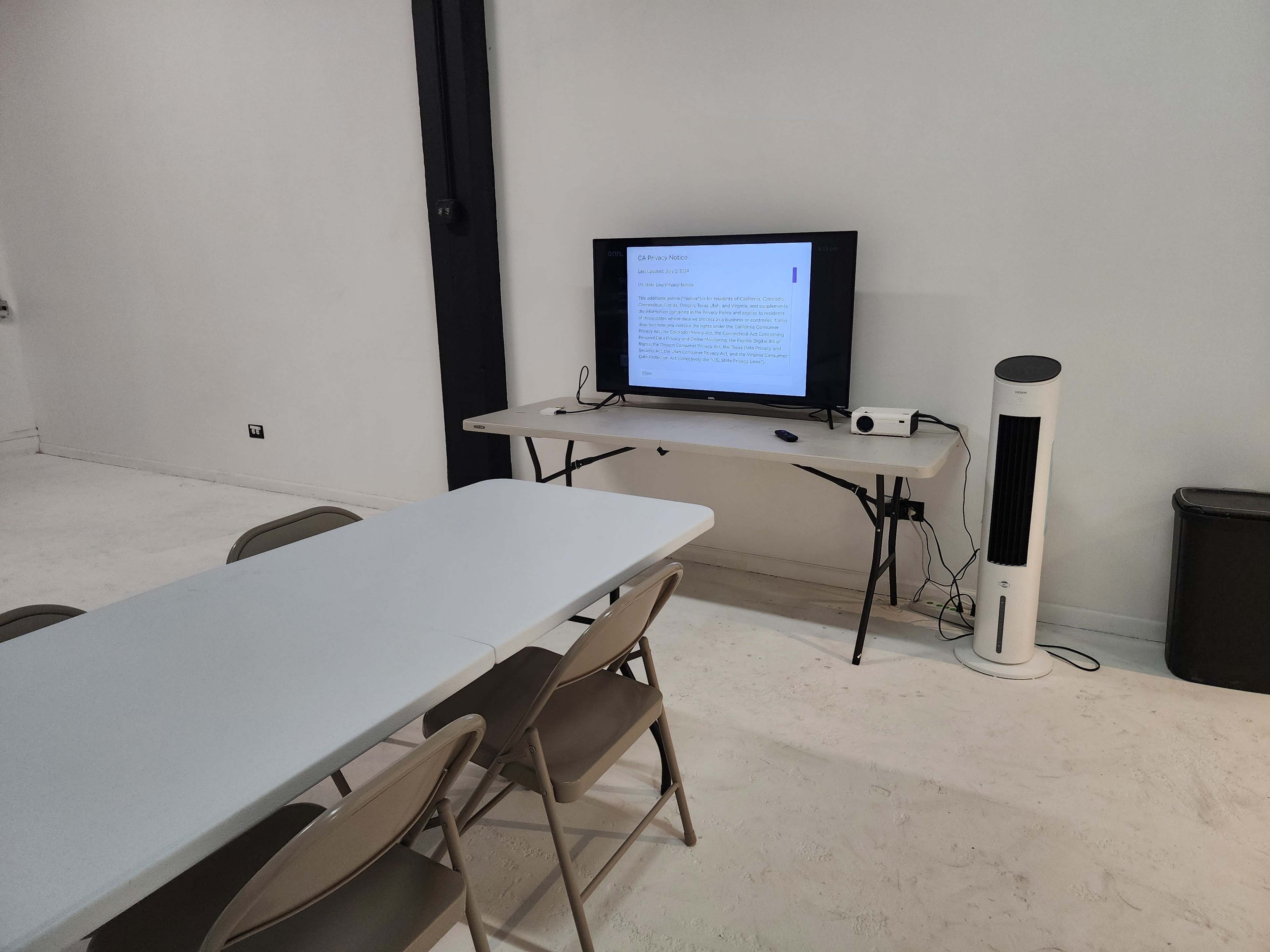 A room with a table and chairs set up for a meeting, featuring a television displaying a presentation and a fan nearby.