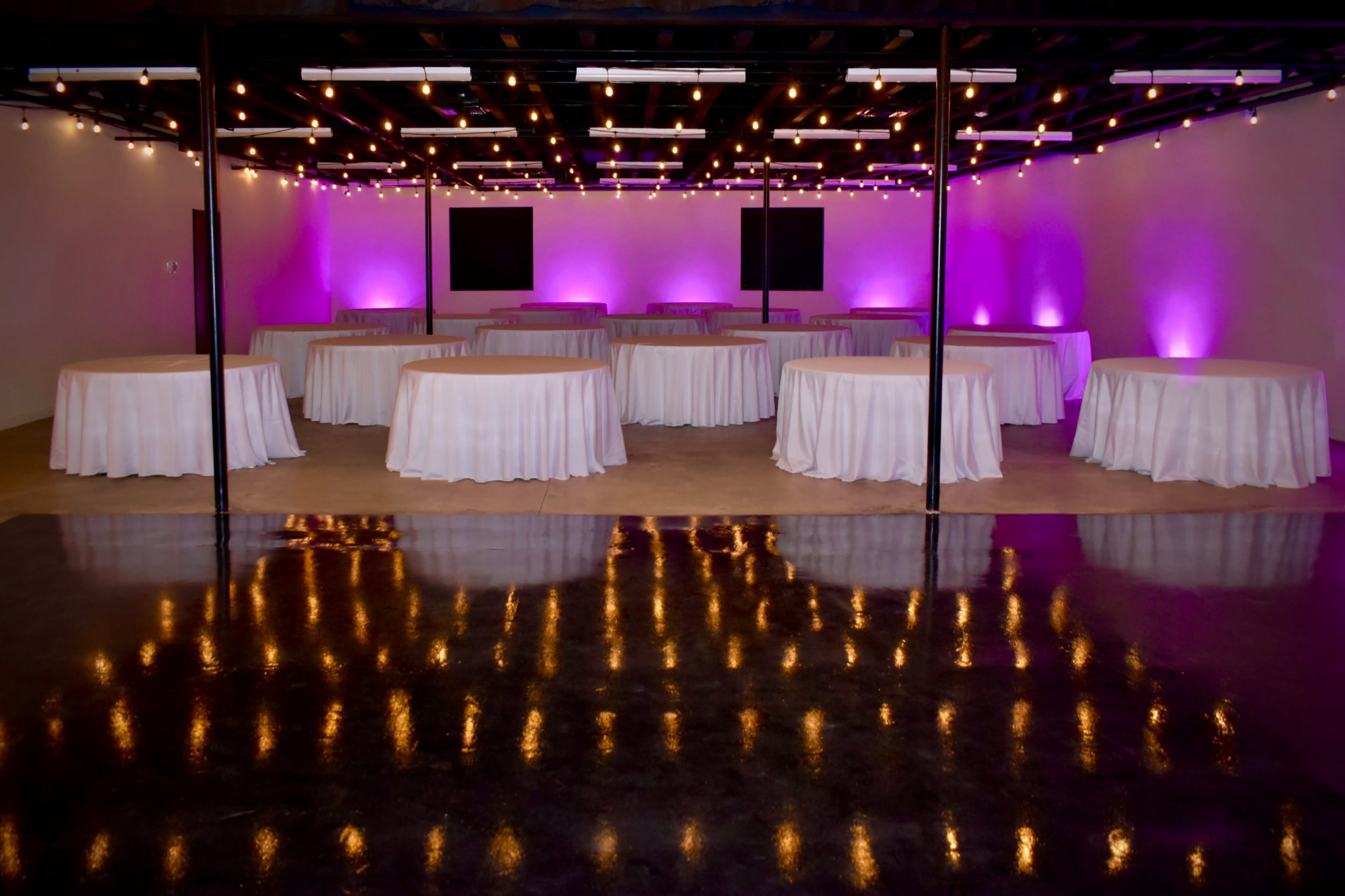 A spacious event venue features several round tables covered with white tablecloths, illuminated by purple and warm overhead lights reflecting off the polished floor.