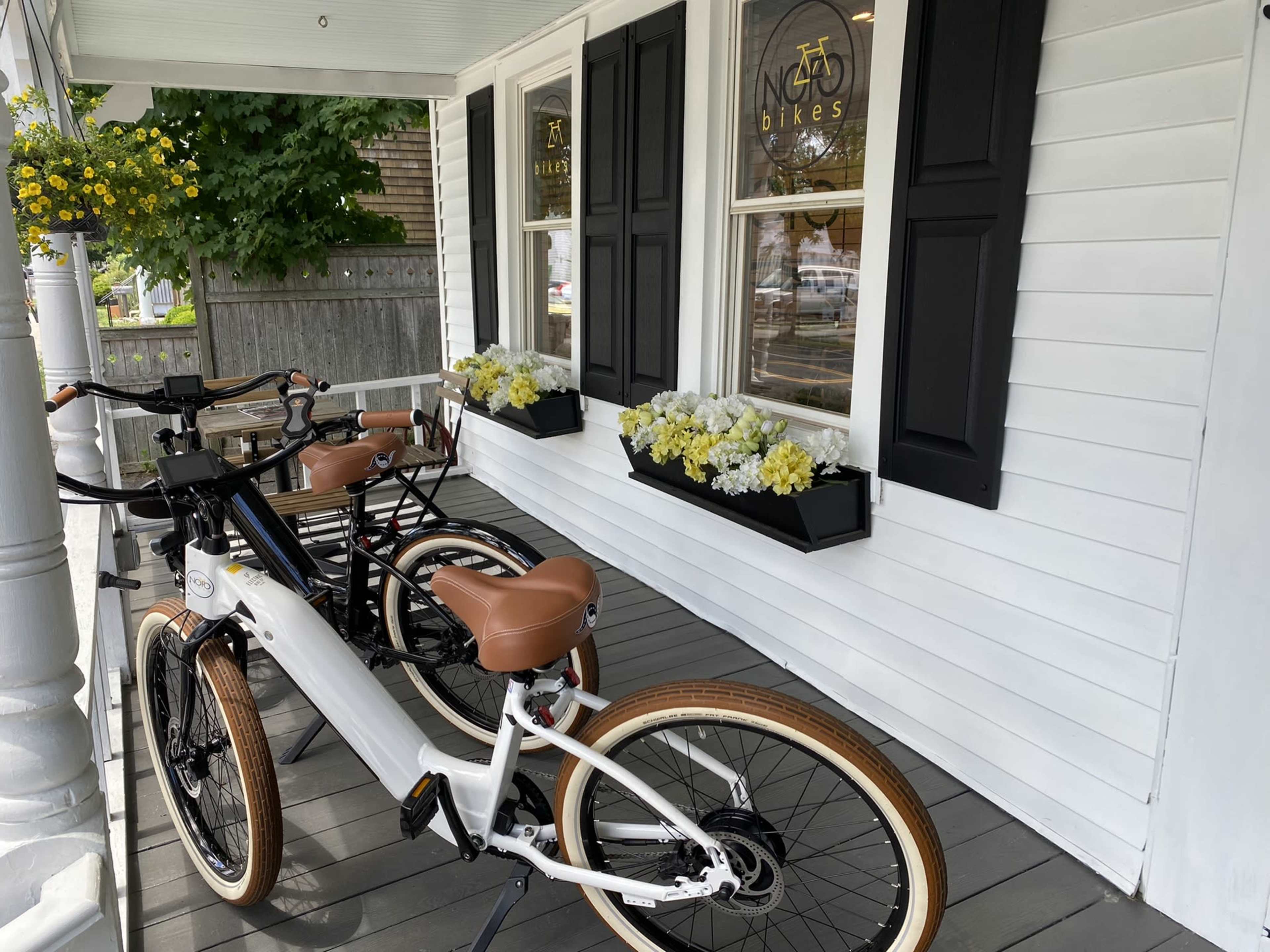 Two bicycles are parked on a porch in front of a shop with flower boxes under the windows.