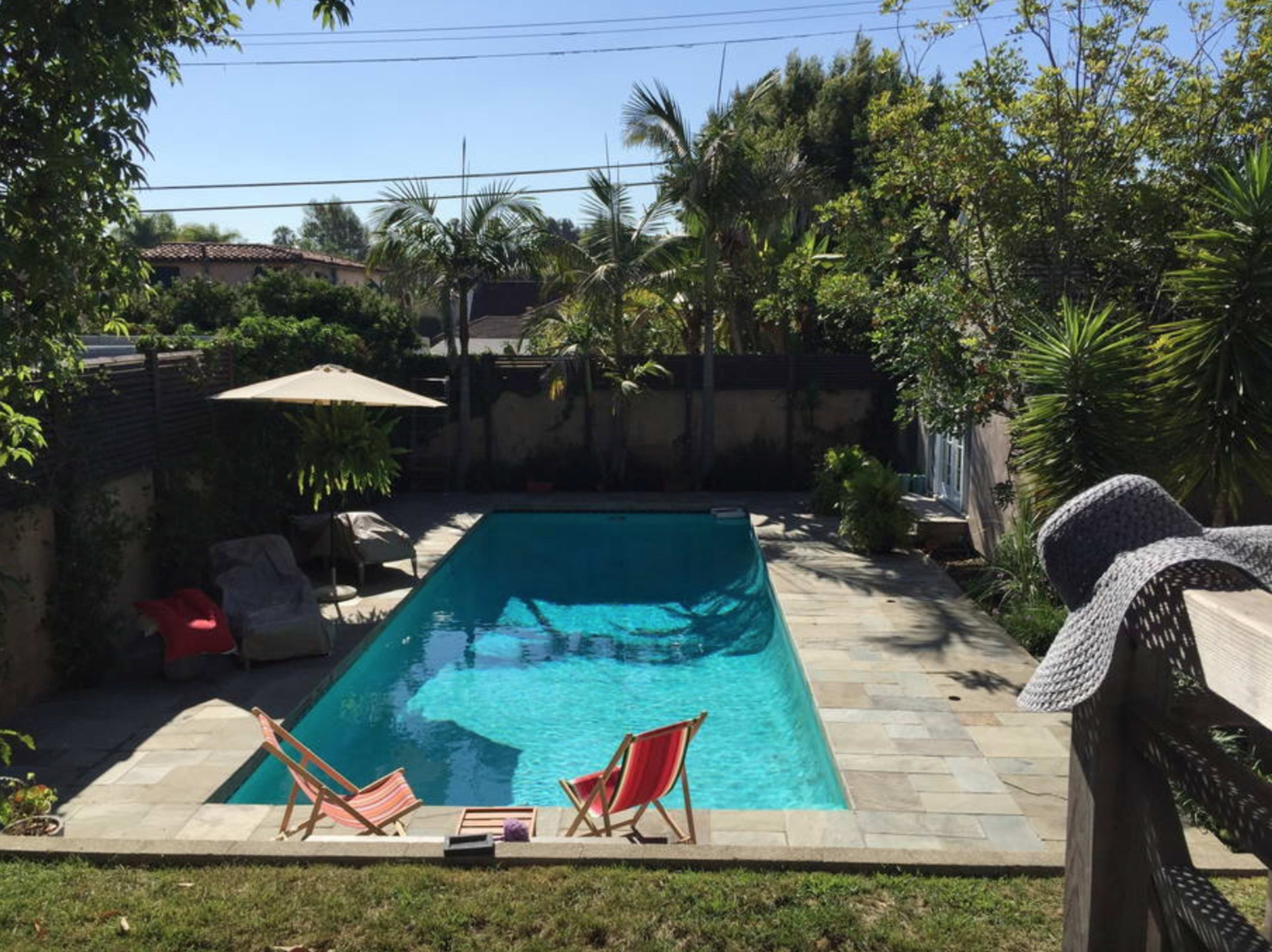 The image shows a rectangular swimming pool surrounded by greenery and lounge chairs on a stone deck.