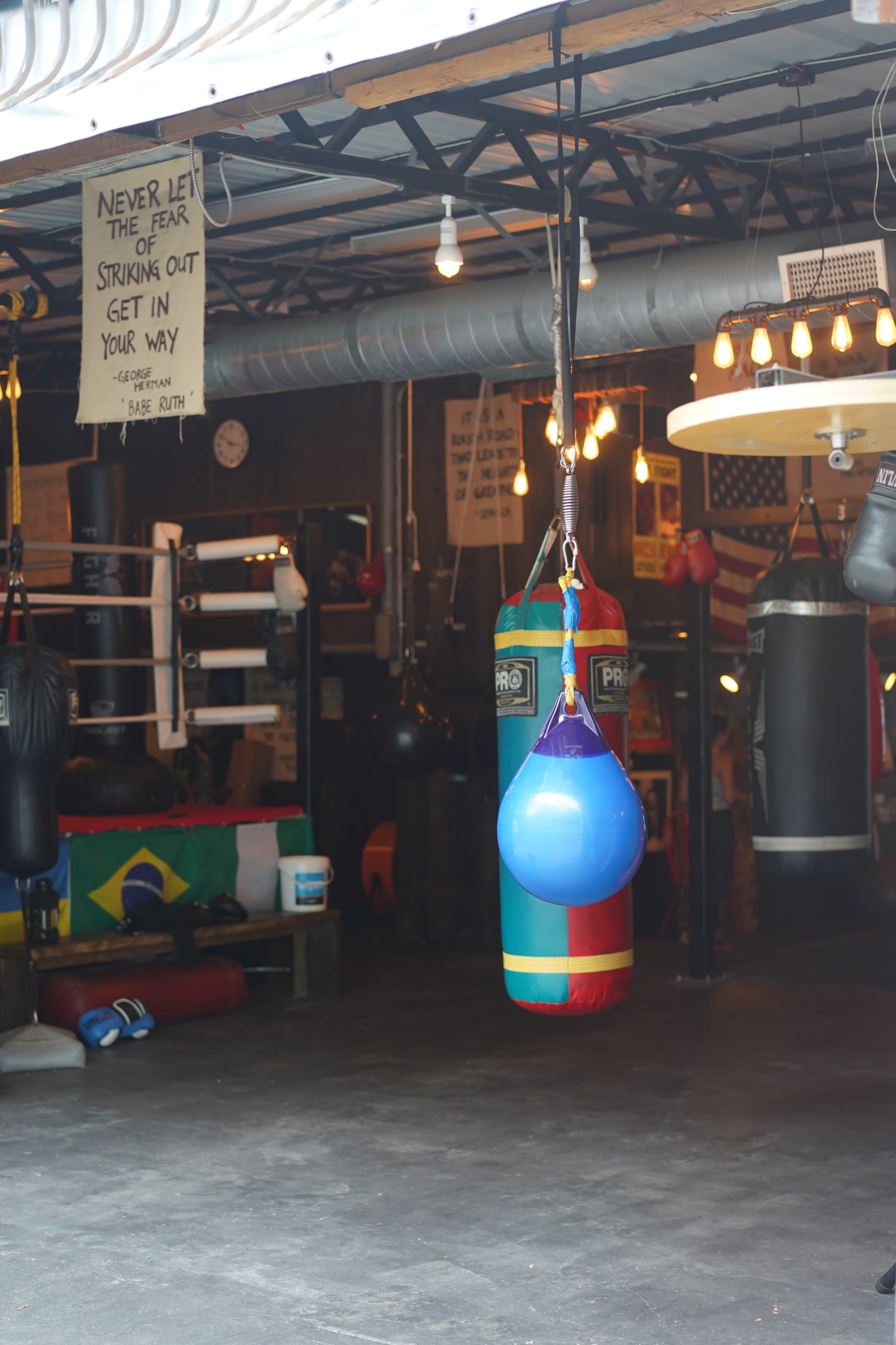 A boxing gym with various hanging bags, motivational signs, and a ring in the background.