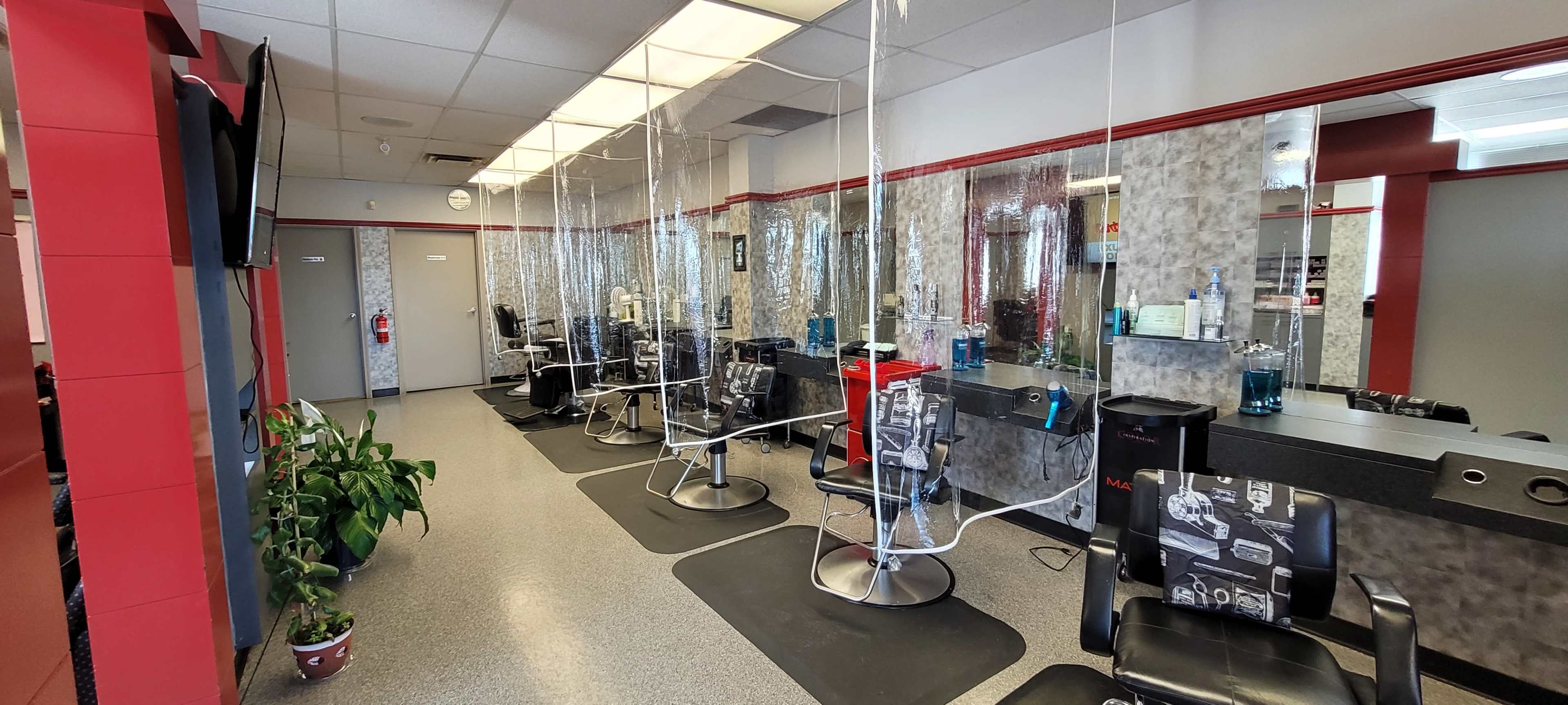 A hair salon interior features several styling stations with clear plastic dividers and black barber chairs arranged along a mirrored wall.