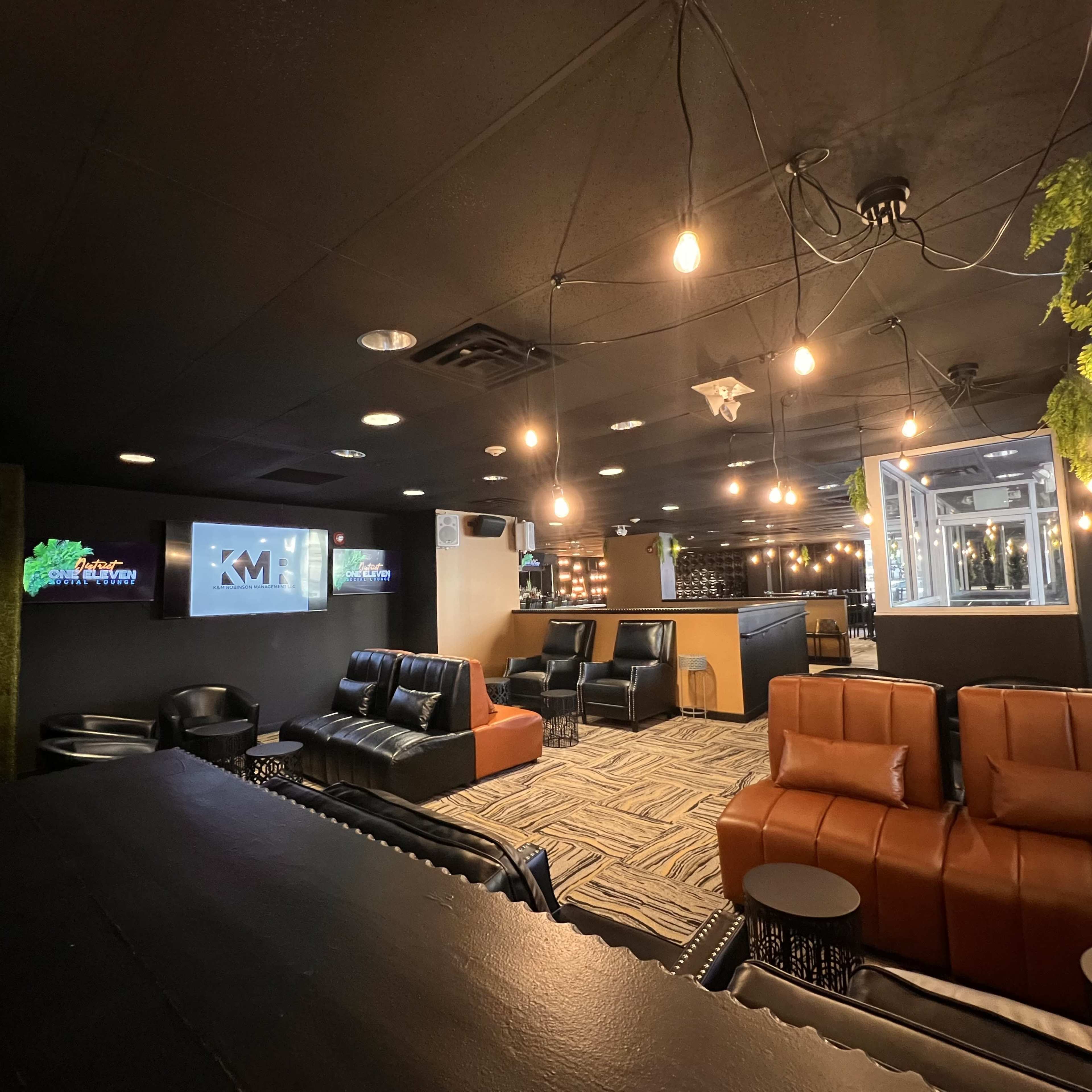 The image shows a modern lounge area featuring black and brown leather furniture, pendant lights, and multiple screens, with a bar counter visible in the foreground.