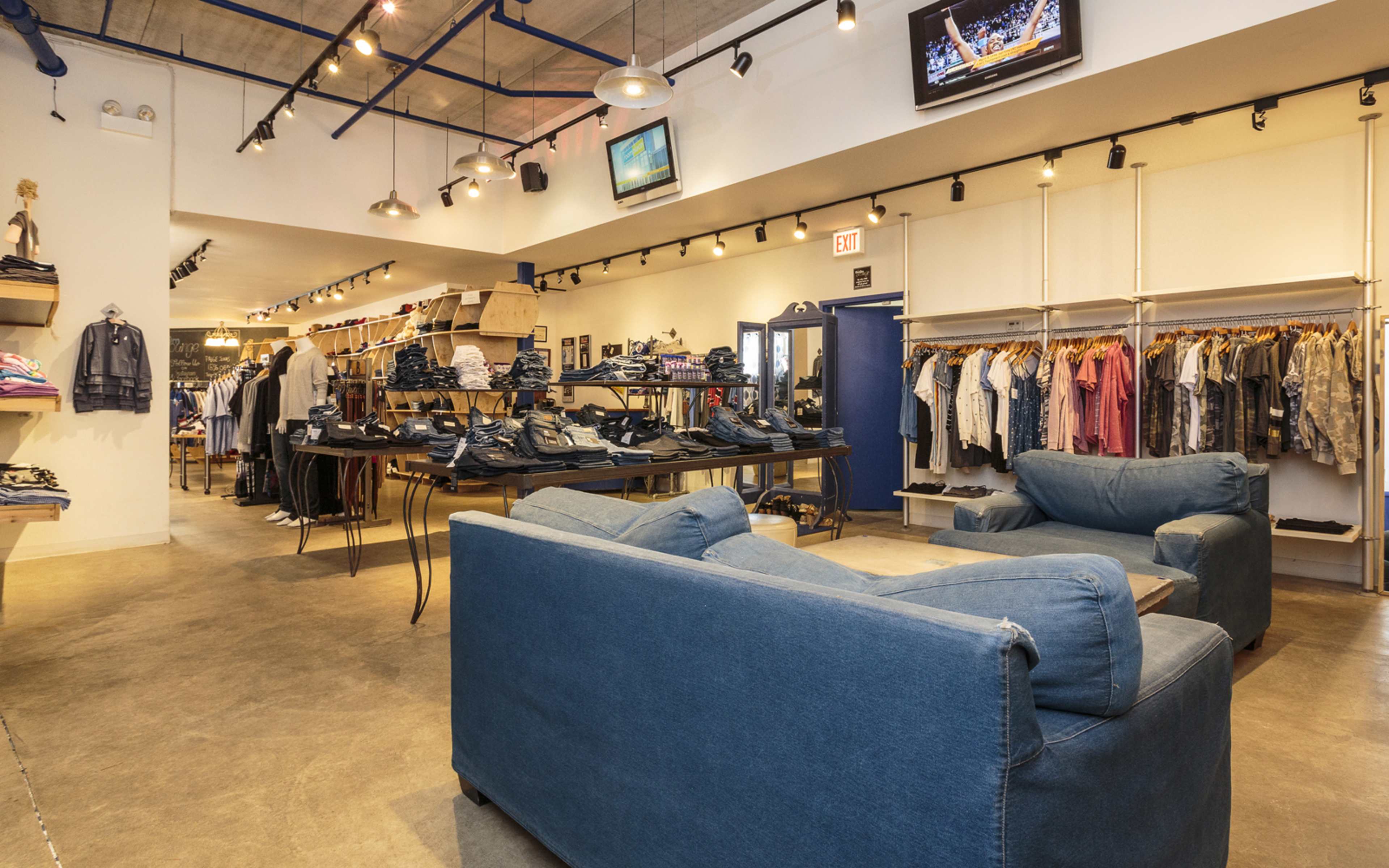The image shows a retail clothing store with a display of clothes on racks, a large table with folded items, and seating areas with blue couches.