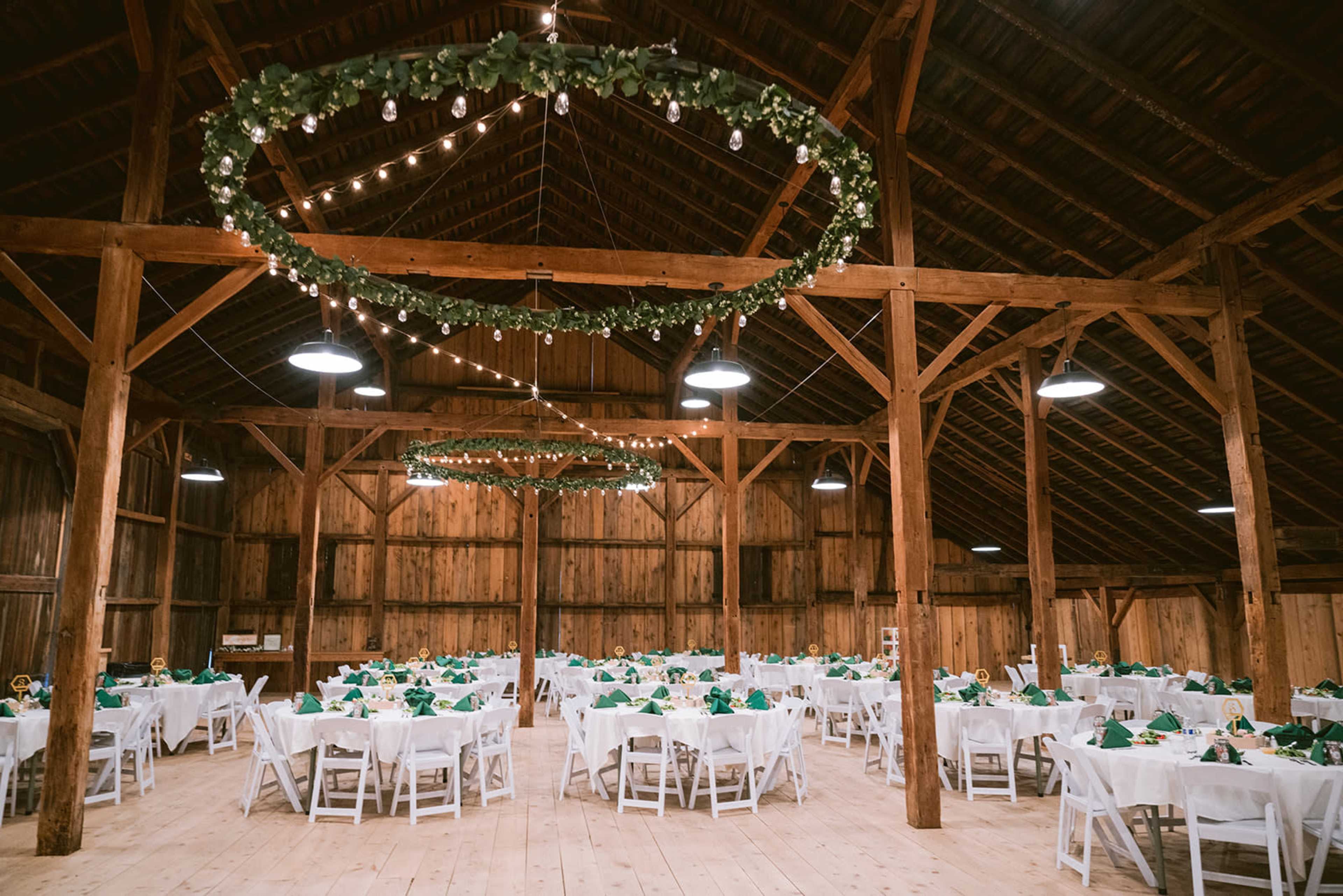 The scene shows a rustic barn interior set up for a formal event, featuring tables neatly arranged with white tablecloths and green decorations, illuminated by overhead string lights and circular greenery displays.
