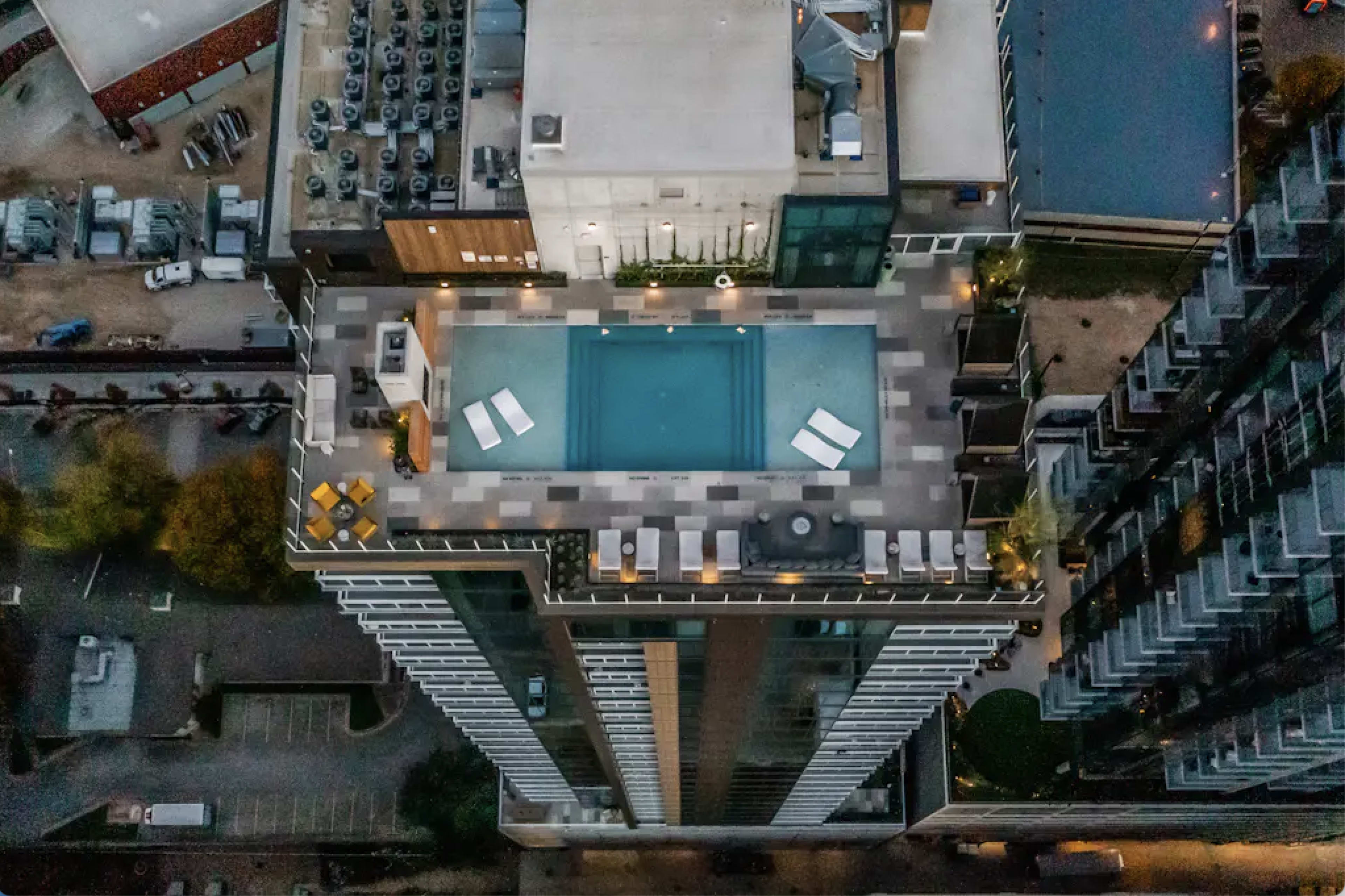 The image shows an aerial view of a high-rise building featuring a rooftop swimming pool surrounded by lounge chairs and a patio area.