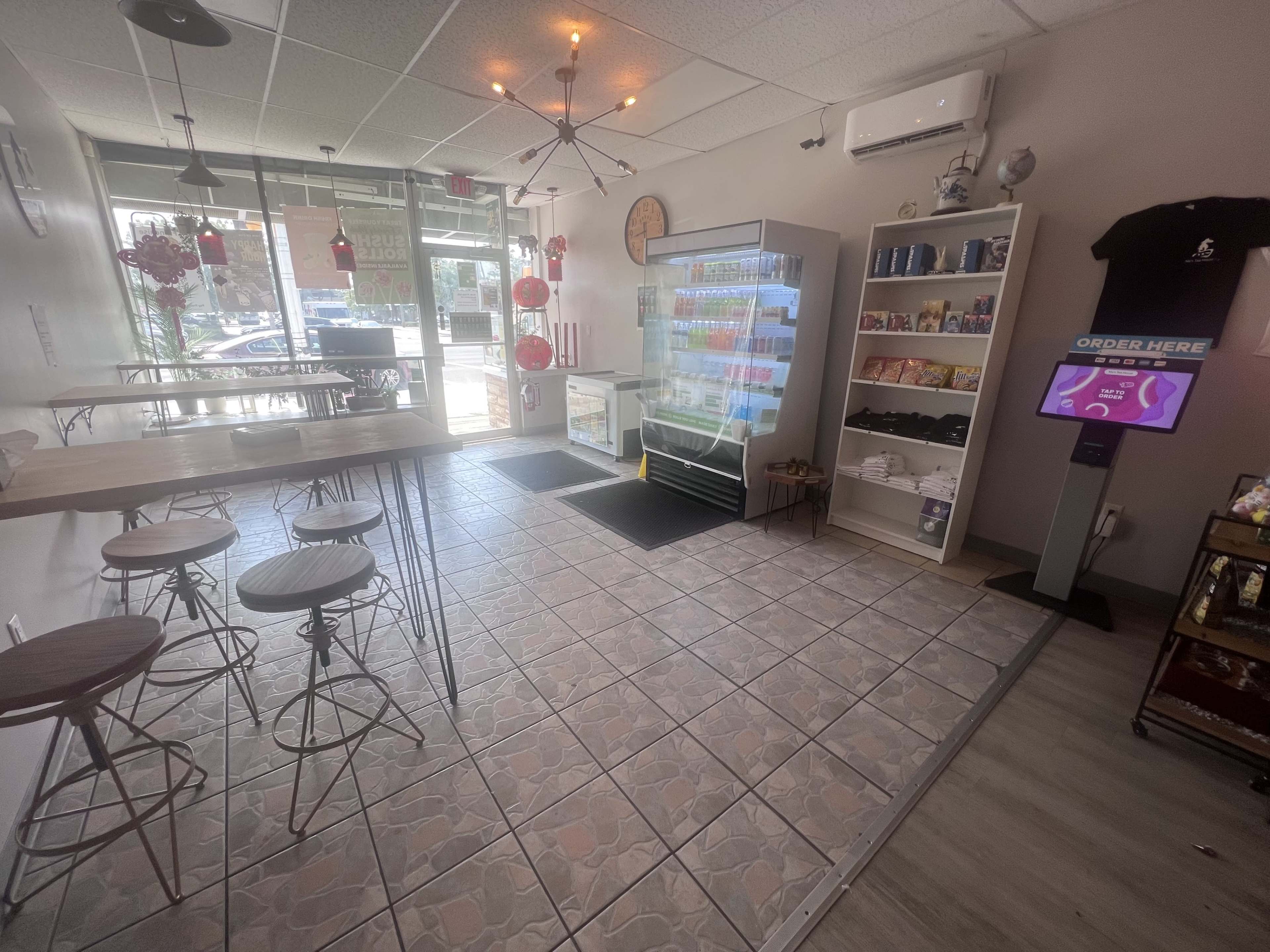 A modern café interior with seating on the left, a display fridge in the center, and a digital ordering kiosk to the right.