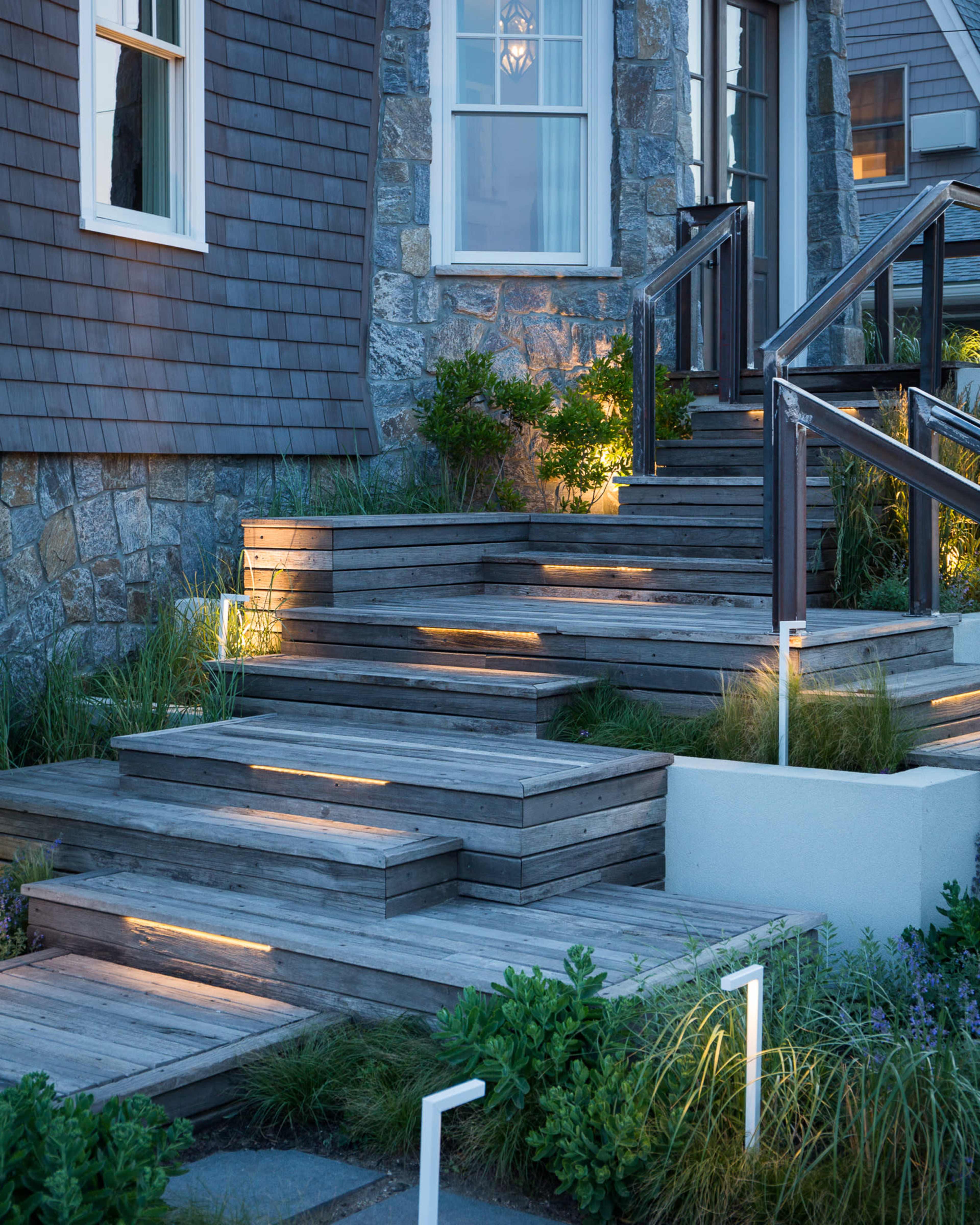 A set of wooden steps leading up to a house, with landscape lights illuminating the edges and grass surrounding the pathway.