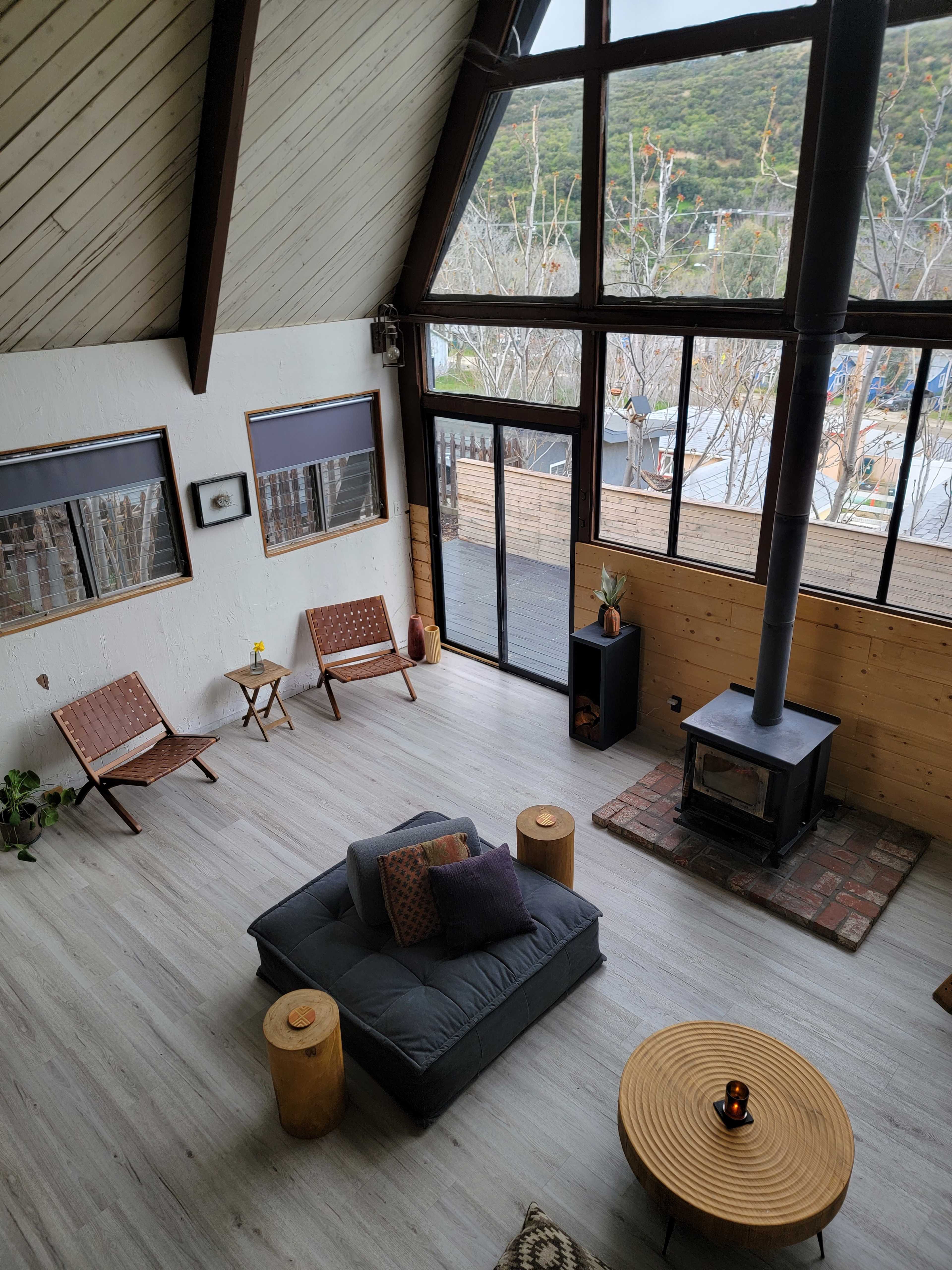 The image shows a spacious living room with a wood-burning stove, minimalist furniture, and large windows letting in natural light.