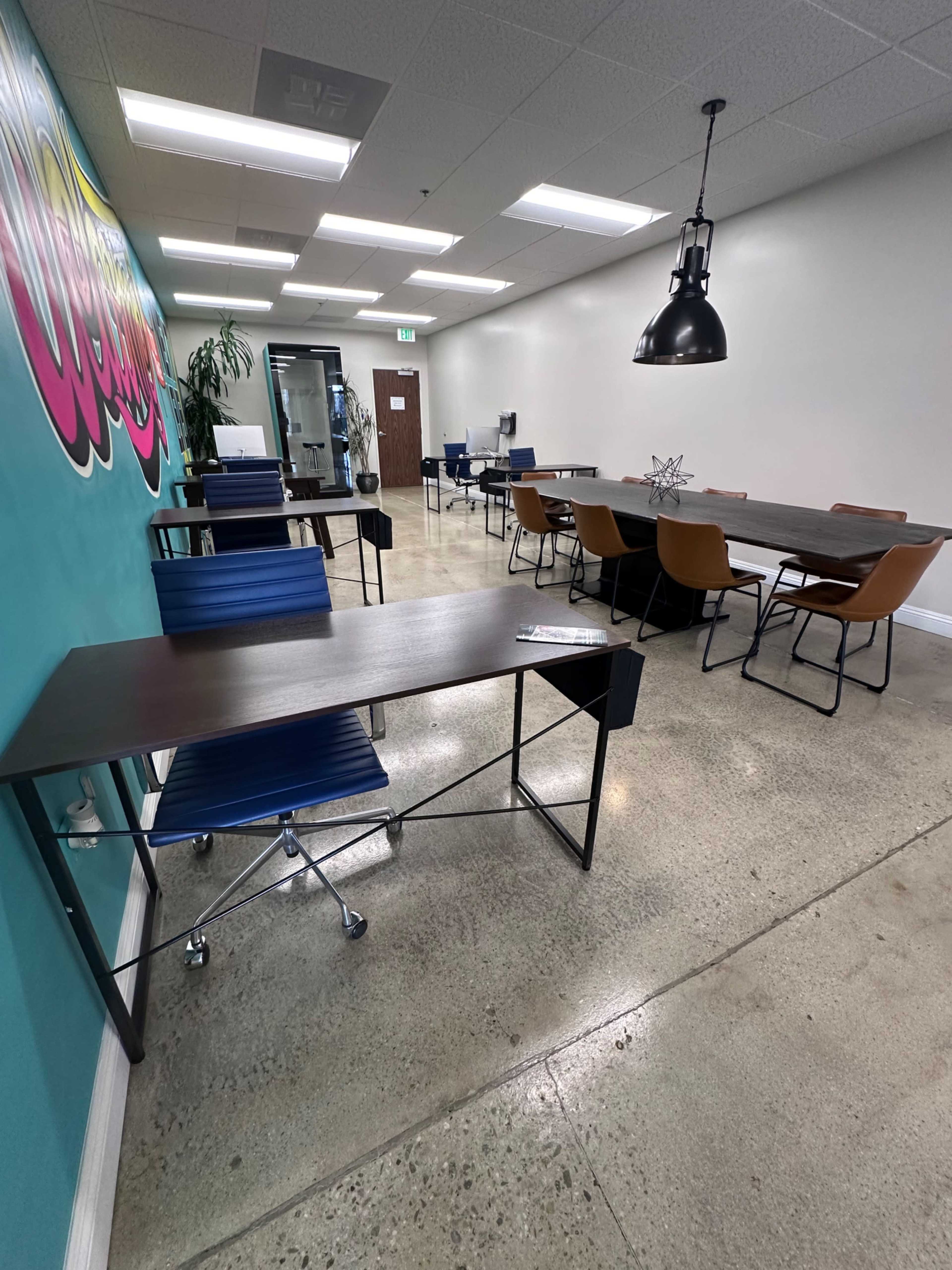 A modern office space with multiple workstations, including desks and chairs, along with a colorful mural on one wall.