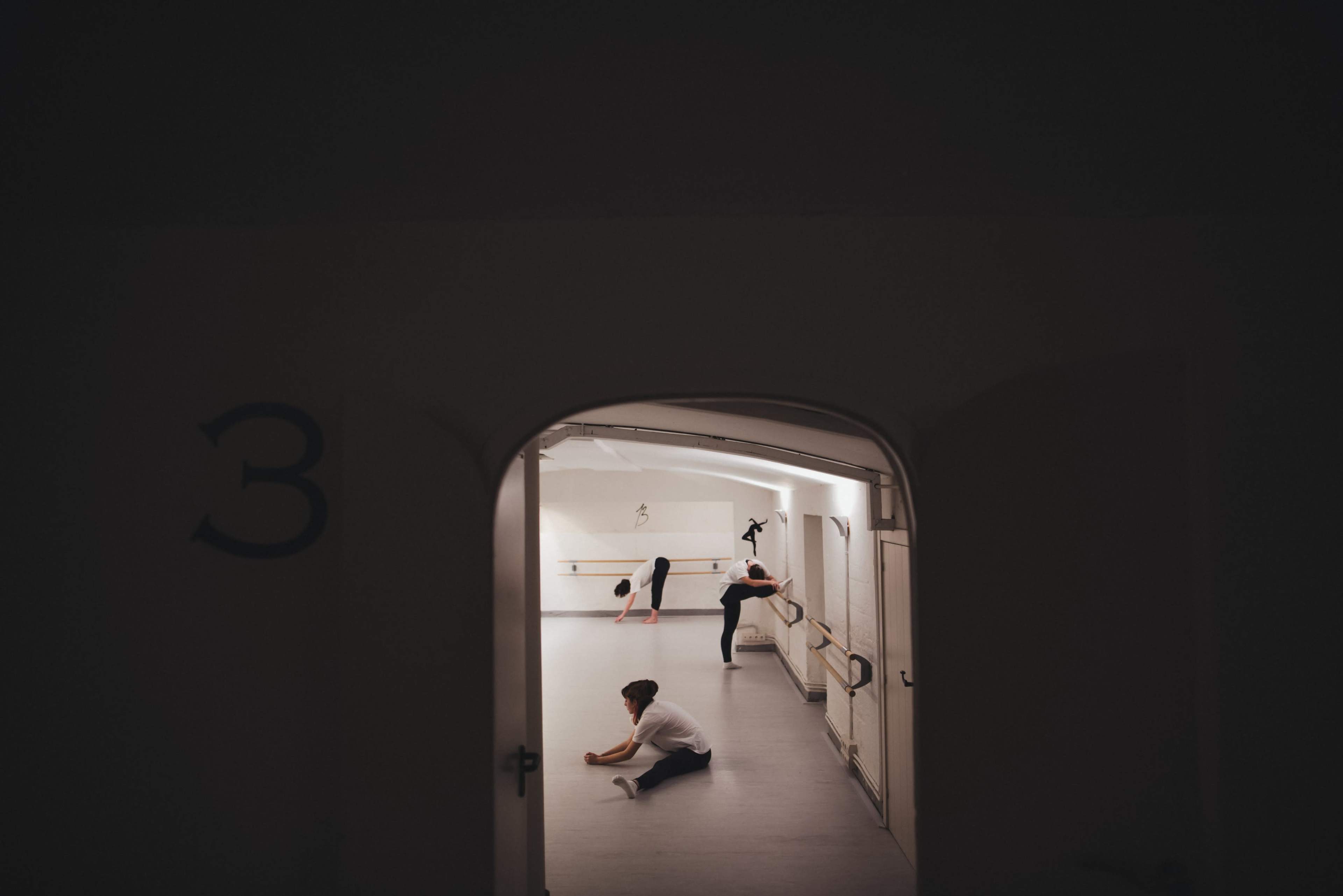 Two dancers stretch and practice in a well-lit studio, with a third dancer in the background near a ballet barre.