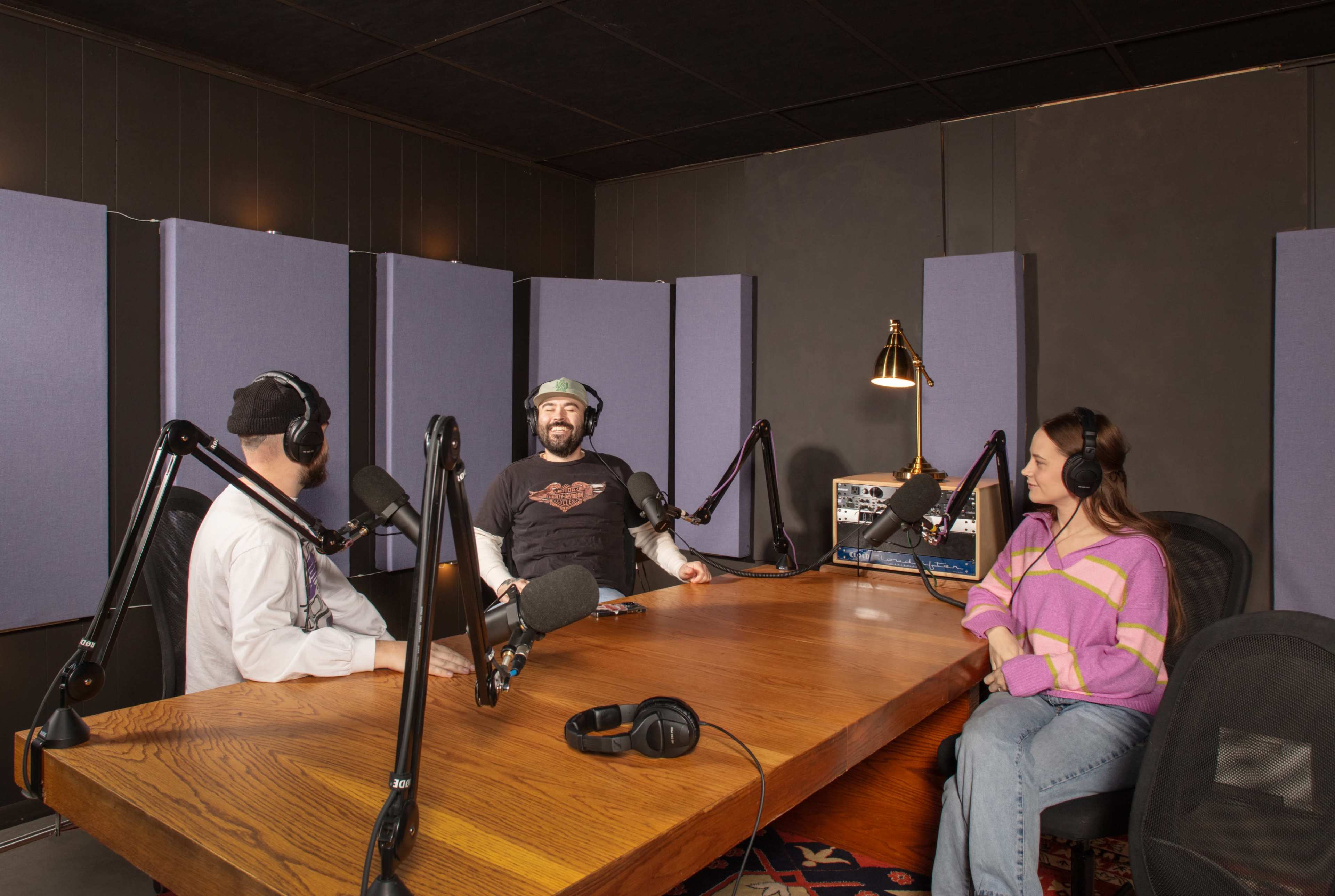 Three people are seated at a wooden table in a podcast studio, equipped with microphones and soundproofing panels.