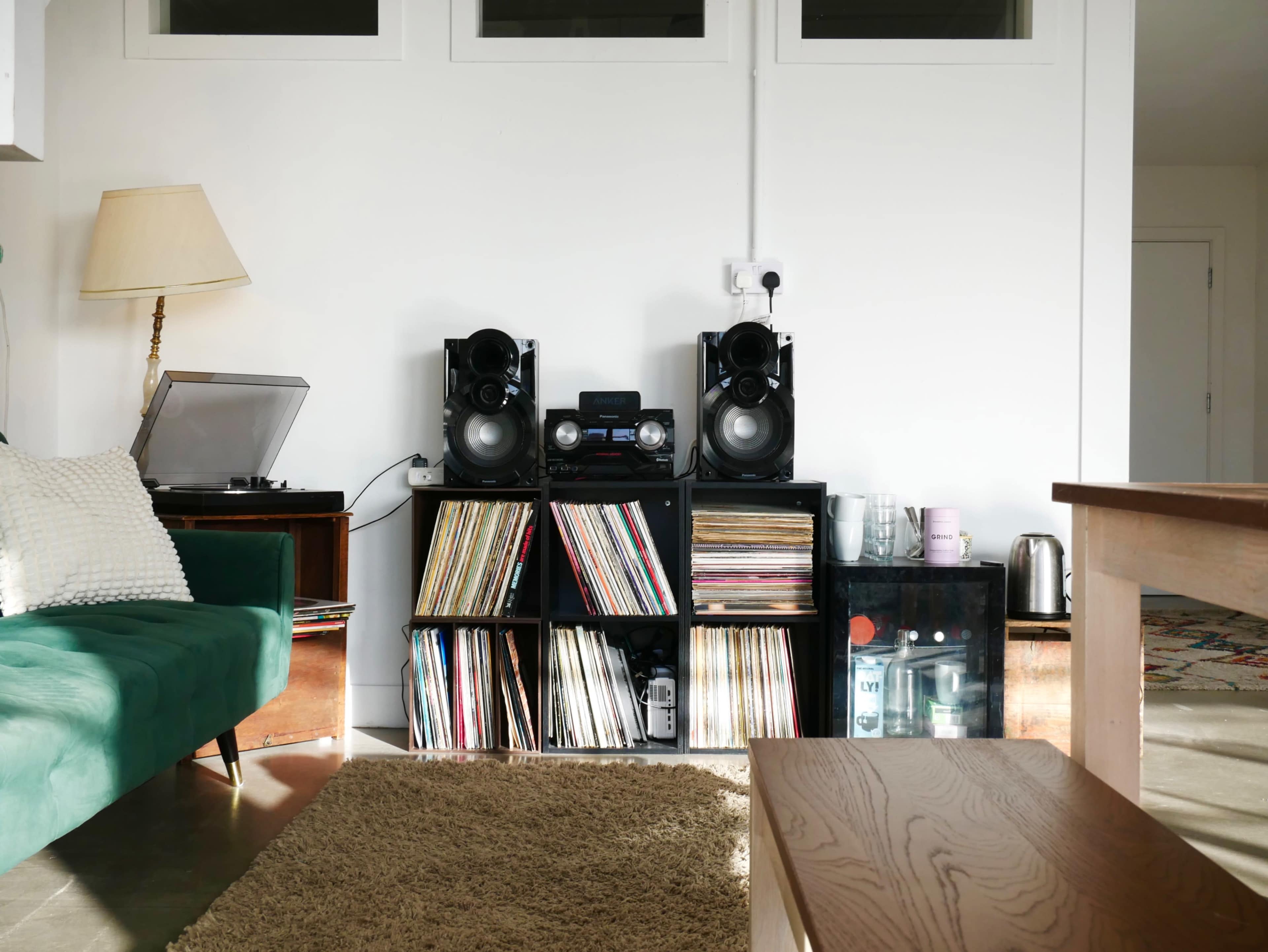 The image shows a living room corner with a record player, two speakers, and a shelf filled with vinyl records.