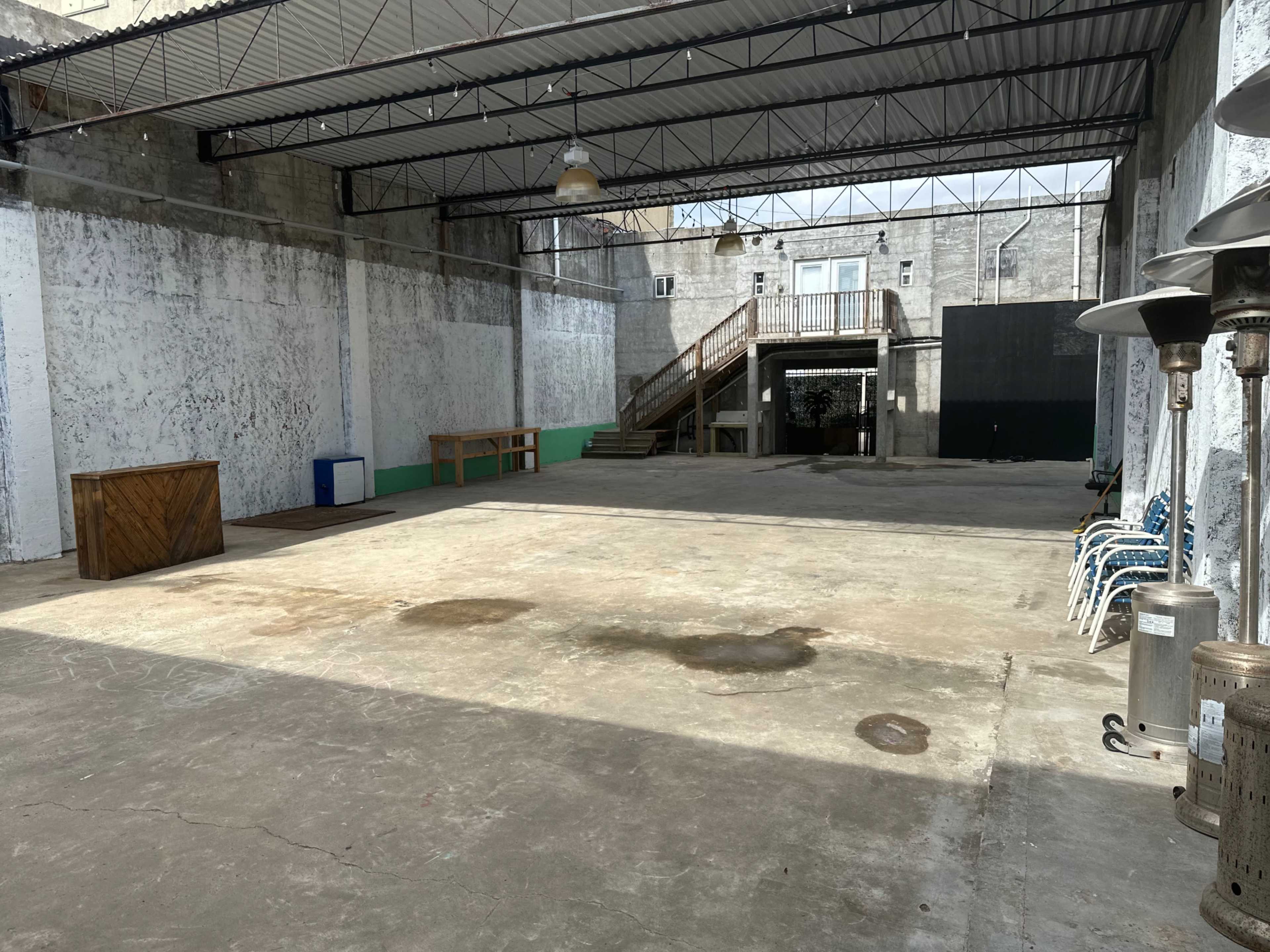 The image shows a spacious, empty indoor area with concrete flooring, high ceilings, and large windows allowing natural light, along with some furniture and heating elements along the walls.