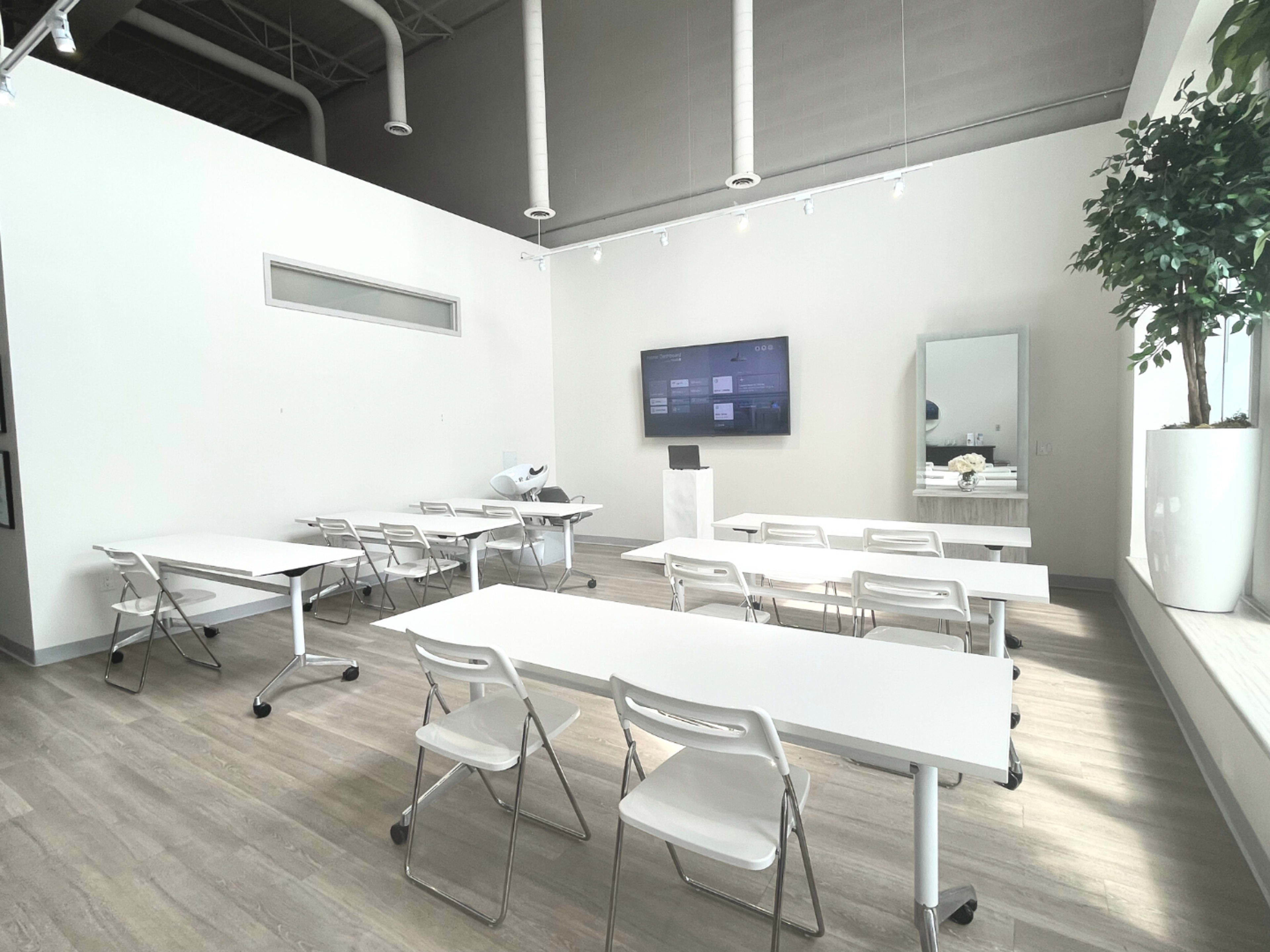 A modern classroom or meeting space with white tables, chairs, a large screen on the wall, and a potted plant in a corner.