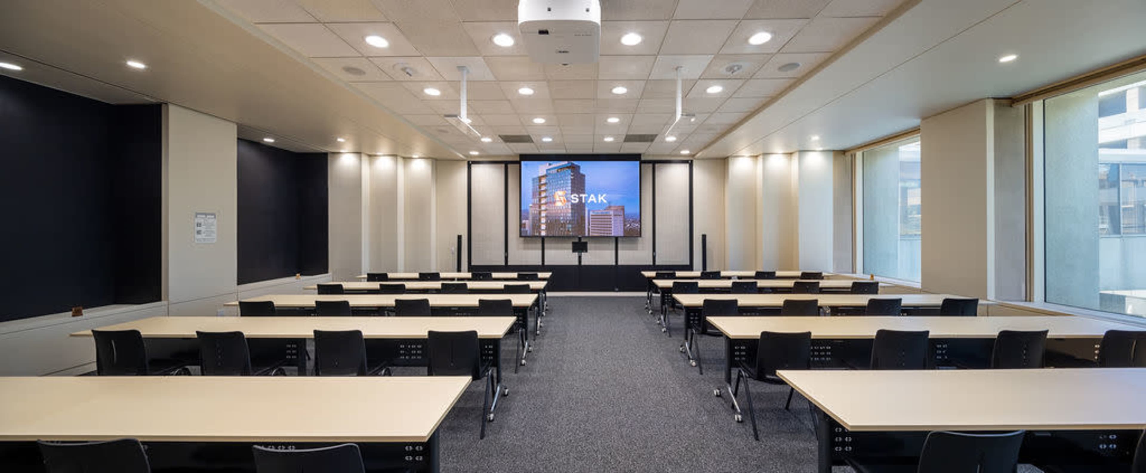 The image shows a modern conference room with multiple rows of tables and chairs facing a large screen.