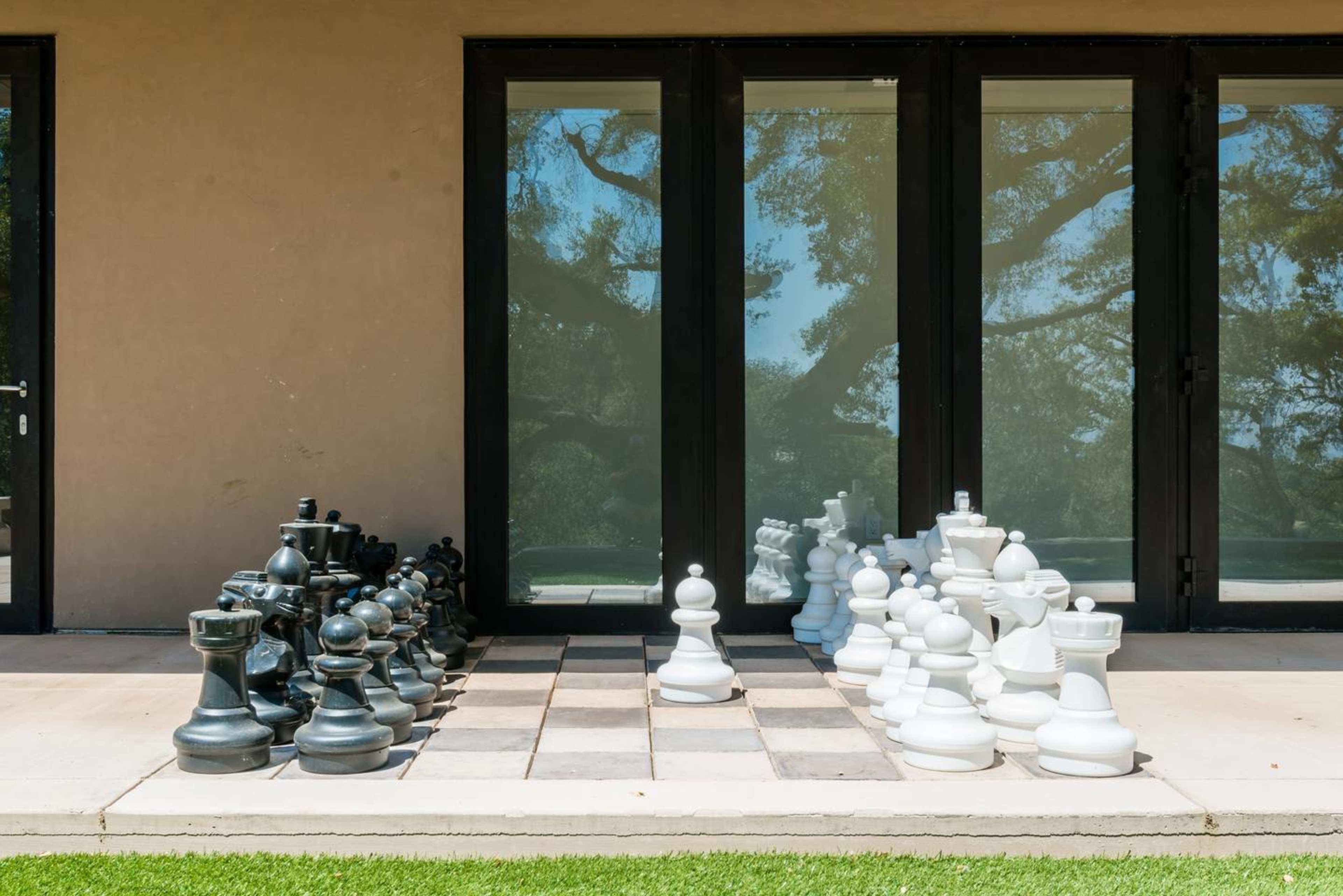 A large outdoor chessboard displays oversized black and white chess pieces arranged for a game in front of glass doors.