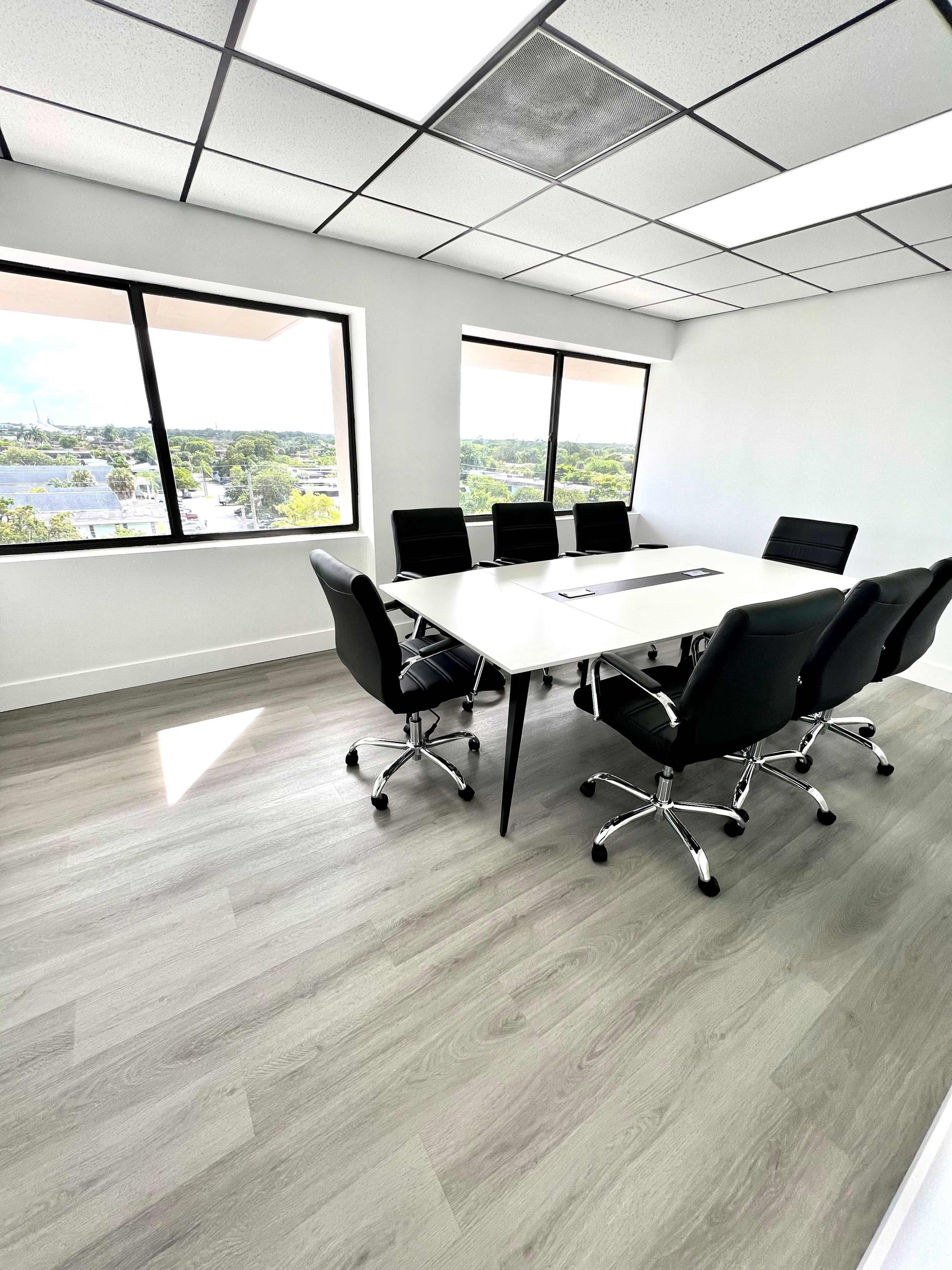 A bright conference room features a large table surrounded by six black chairs and large windows overlooking a cityscape.