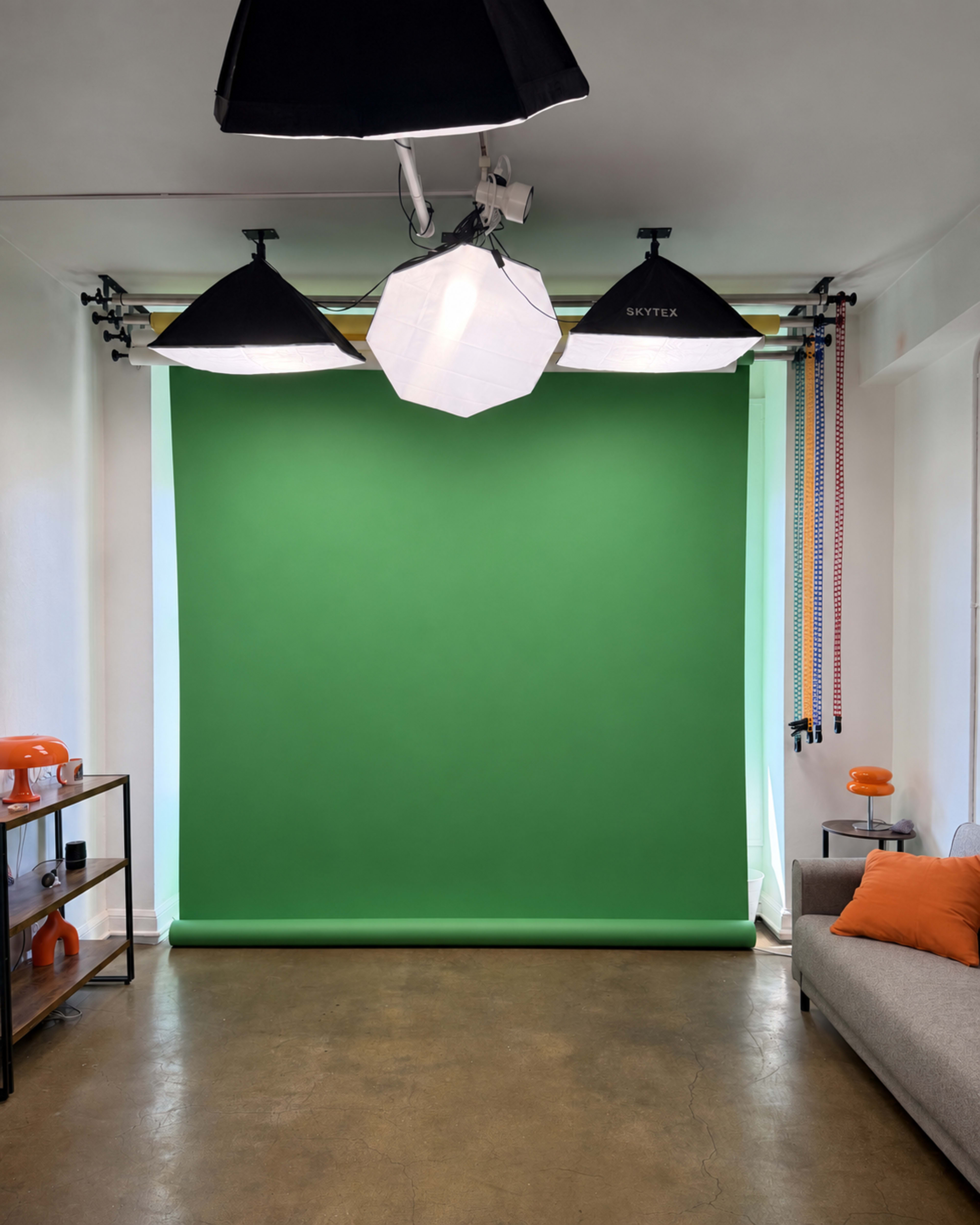 The image shows a studio setup featuring a green backdrop, three lighting fixtures, and a sofa with an orange pillow.