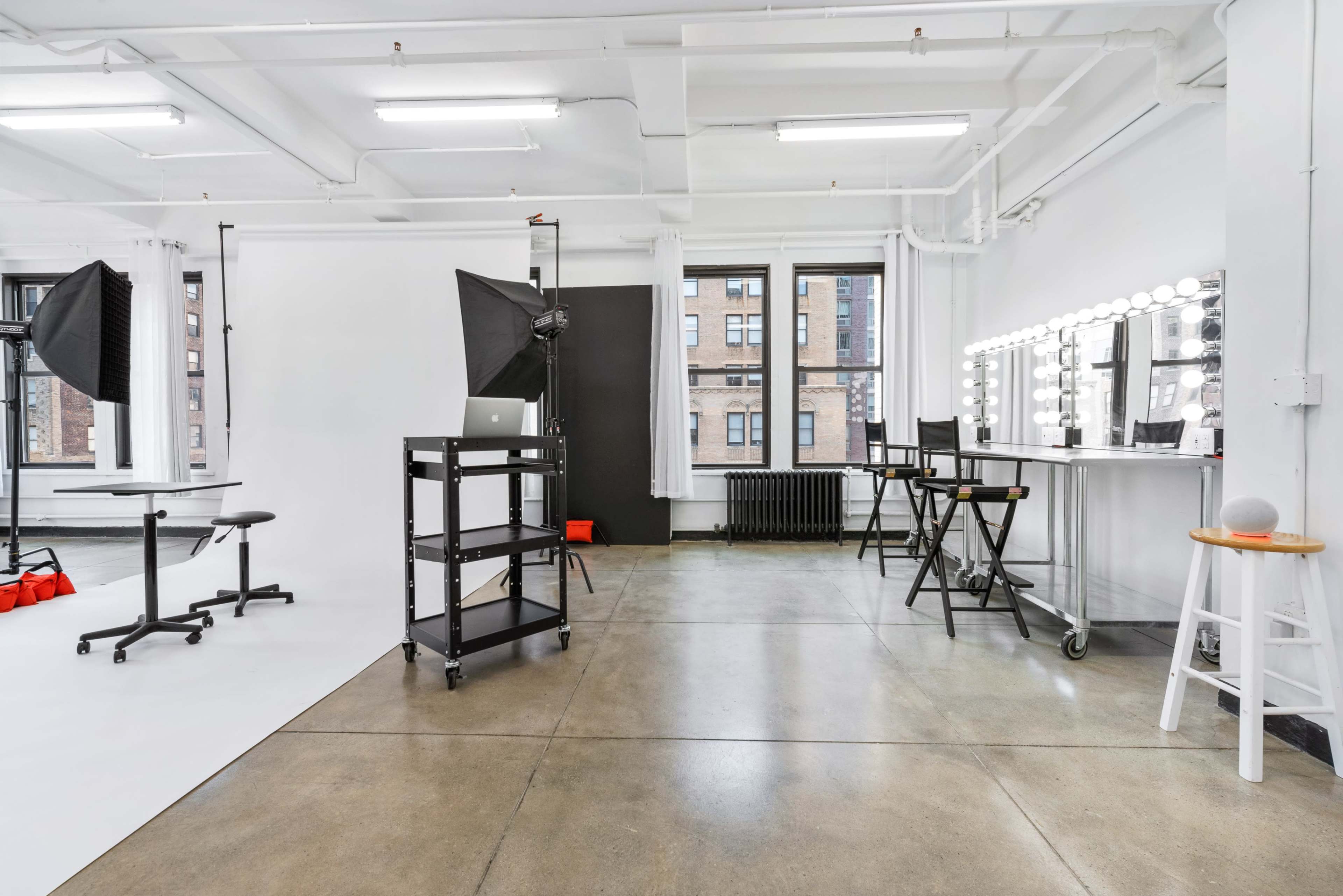 The image shows a bright photography studio equipped with lighting equipment, a large backdrop, and a makeup station.