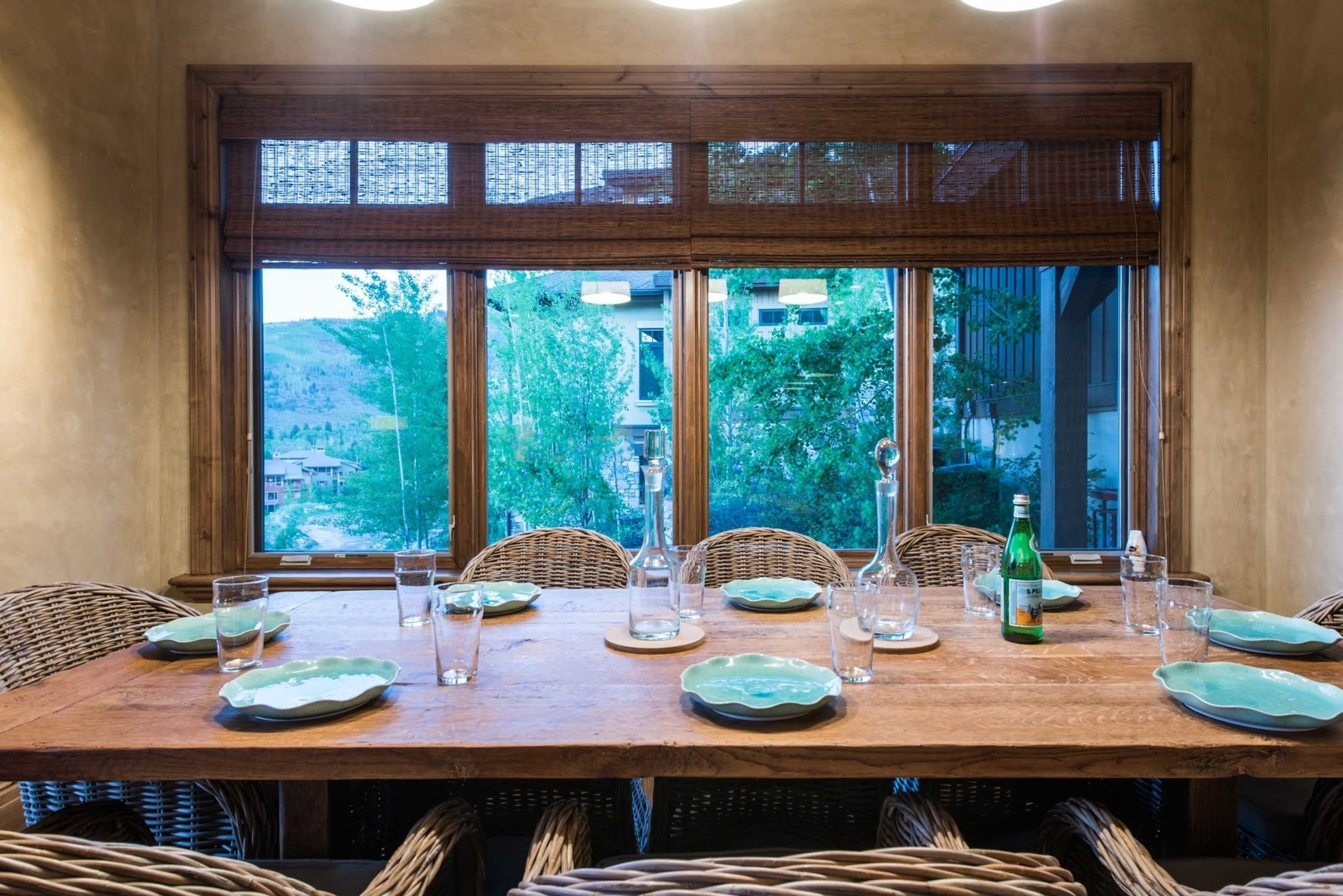 A wooden dining table is set with plates and glasses in a room with large windows overlooking a green landscape.
