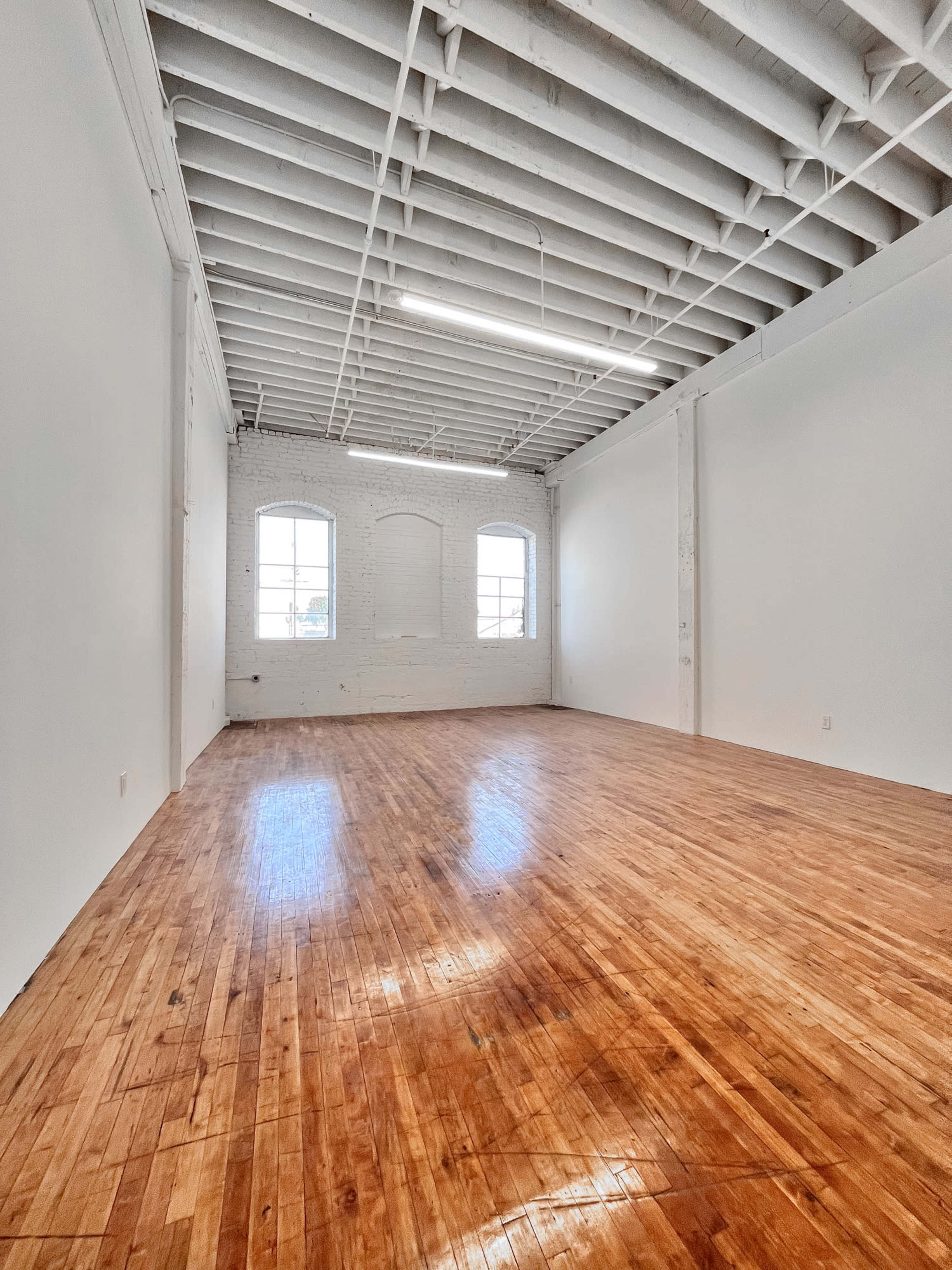 A spacious, empty room with hardwood flooring, white walls, and large windows allowing natural light to enter.