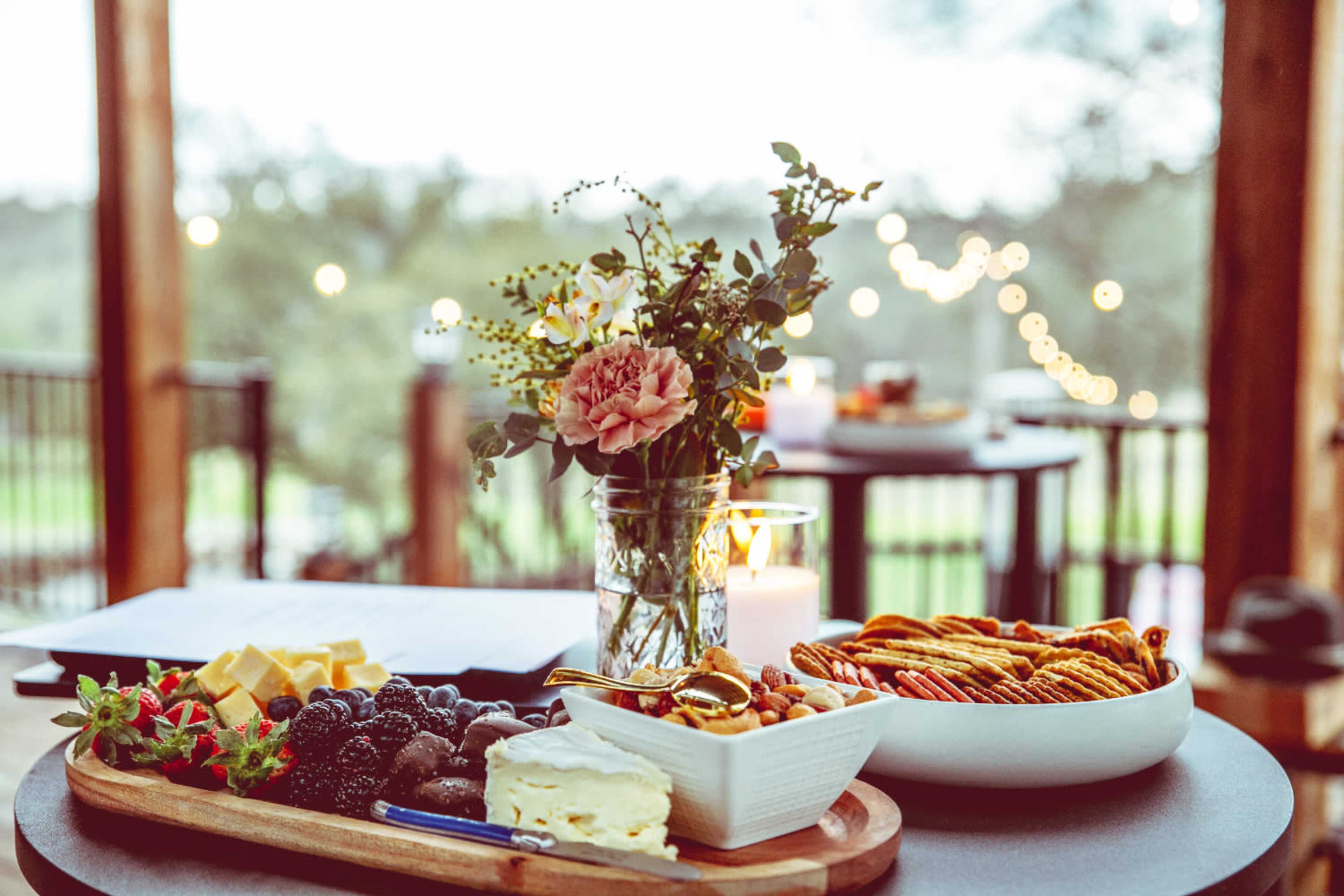 A wooden platter holds assorted cheeses, fruits, nuts, and crackers next to a decorative vase with flowers on a table illuminated by string lights in the background.