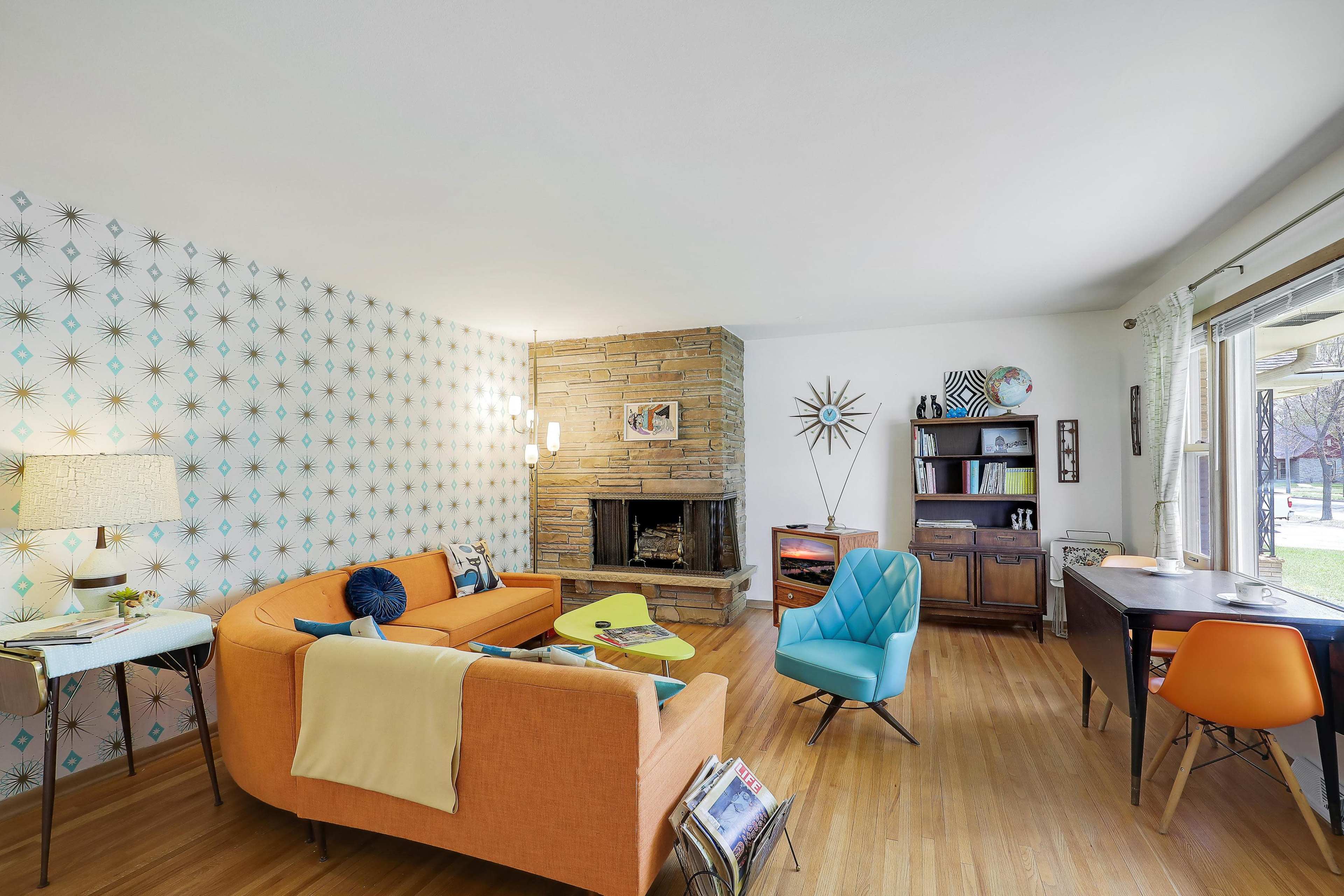 The image shows a living room featuring a fireplace, orange and blue furniture, and wallpaper with a geometric pattern.