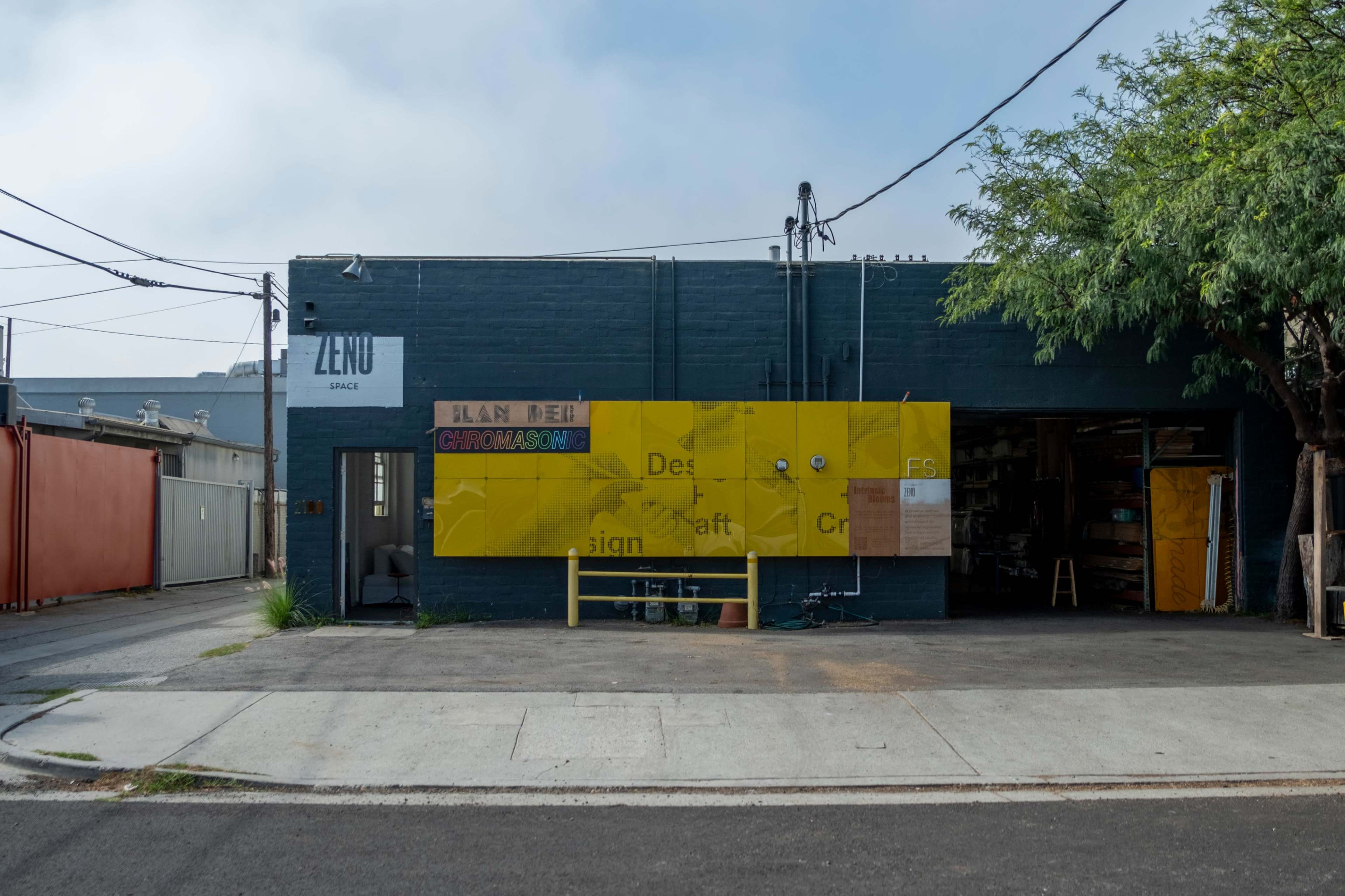 The image shows a gray building with a yellow sign displaying text related to design and craft, set on a street with utility poles and trees nearby.