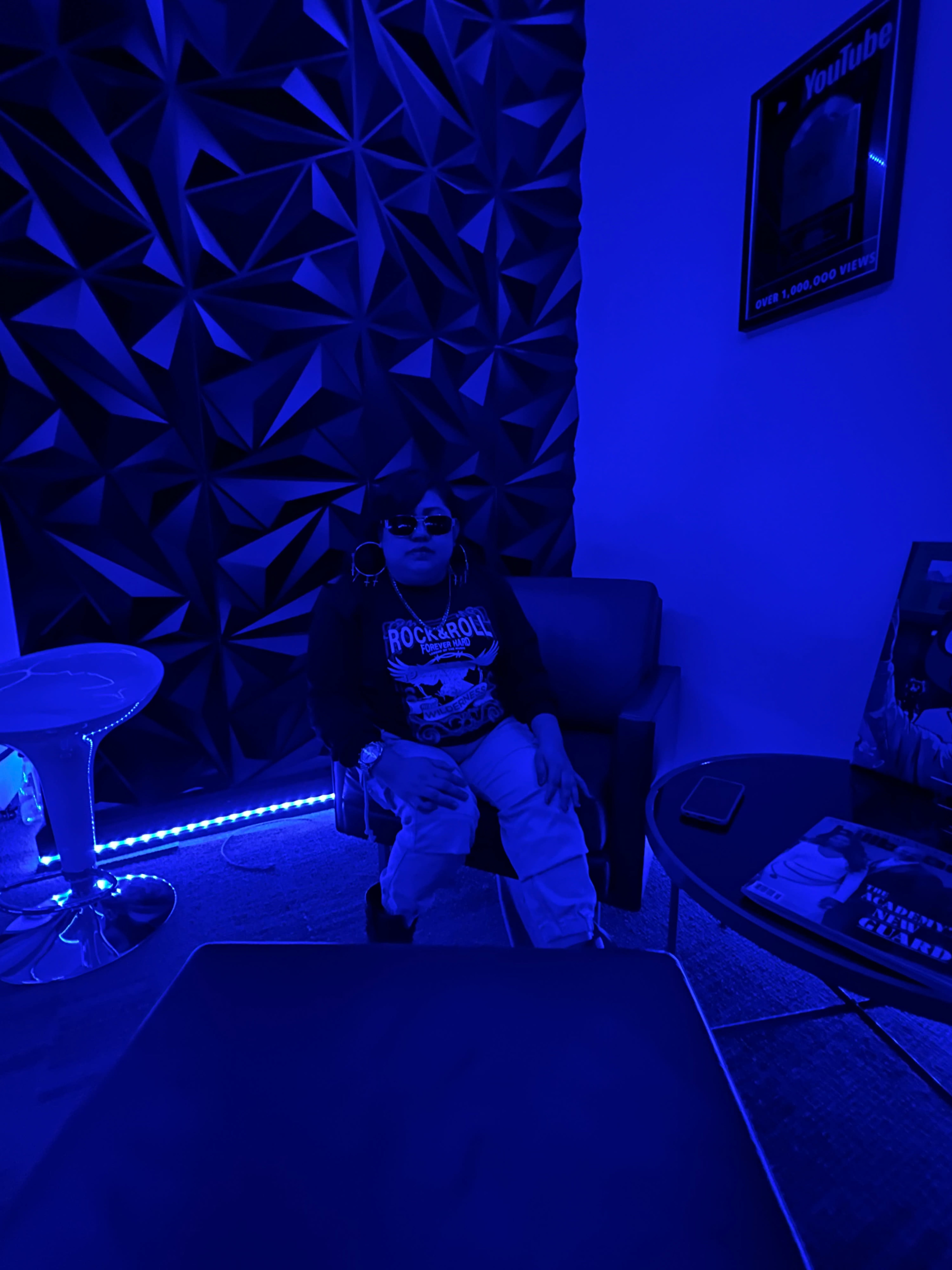 A person wearing sunglasses and a leather jacket sits on a chair in a dimly lit room with blue walls and ambient lighting.