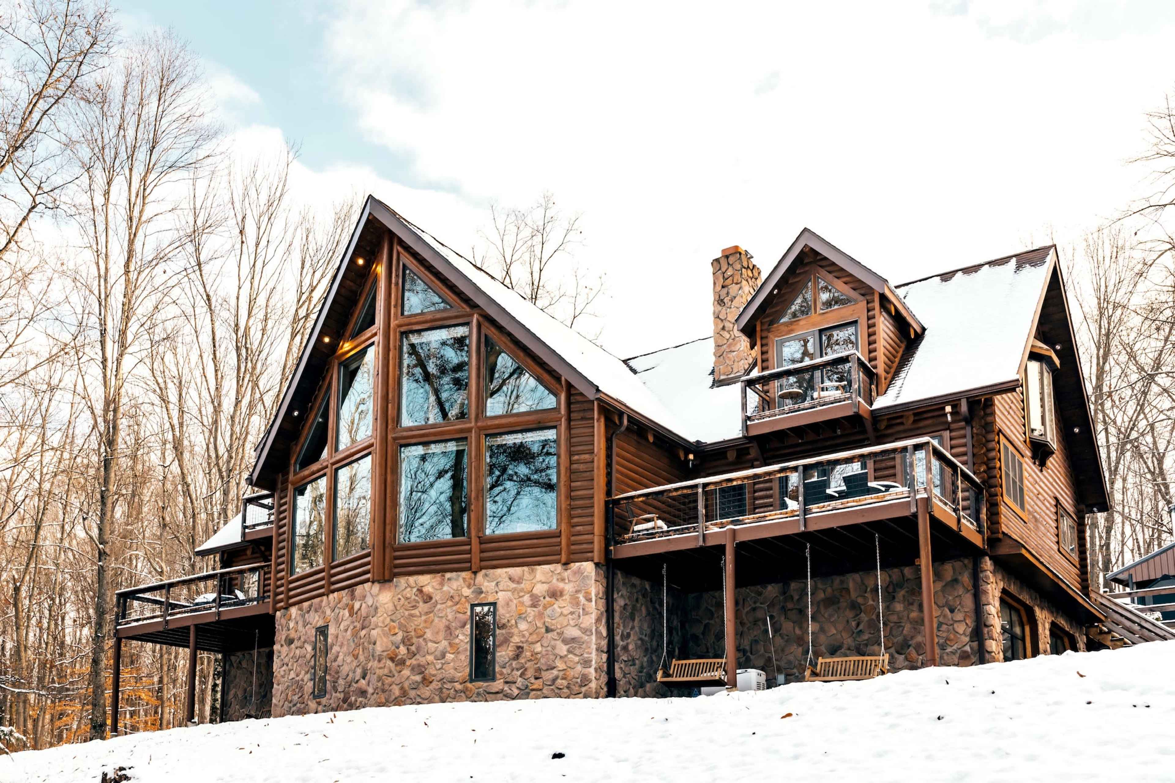 A large log and stone house features multiple large windows and is surrounded by snow-covered ground and trees.