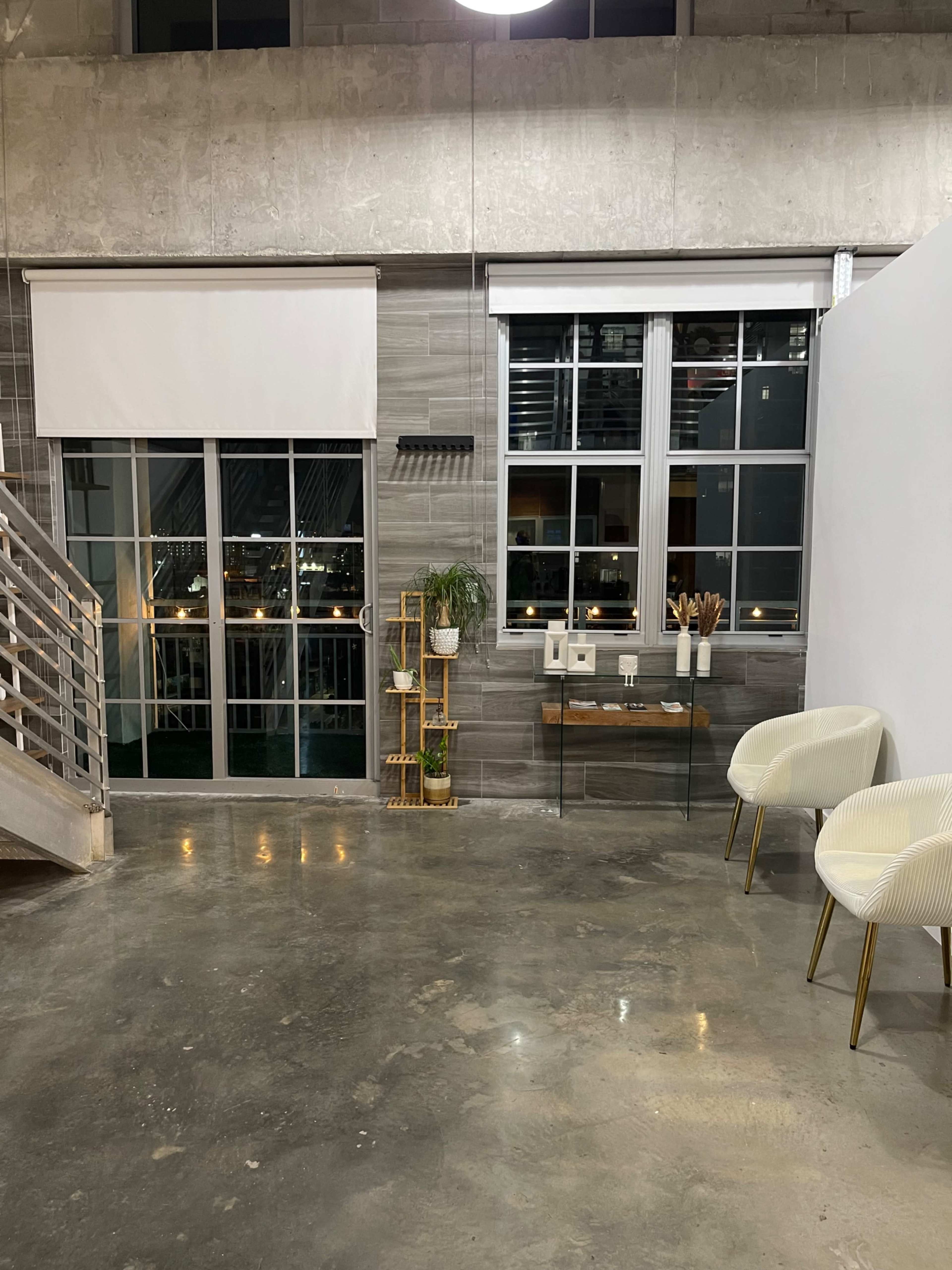 The image shows a modern interior space with large windows, a staircase, a glass table with decor items, and two white chairs.