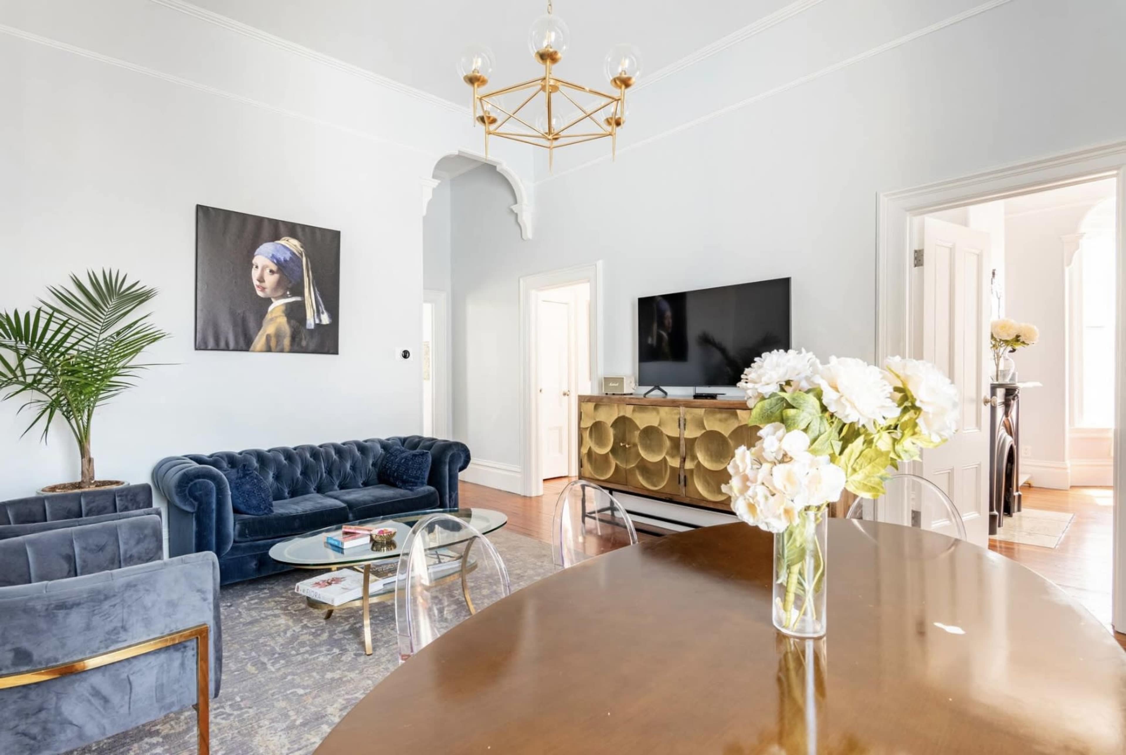 A bright living room featuring a blue velvet sofa, a coffee table, a flat-screen TV on a gold console, and a dining table with a vase of white flowers.