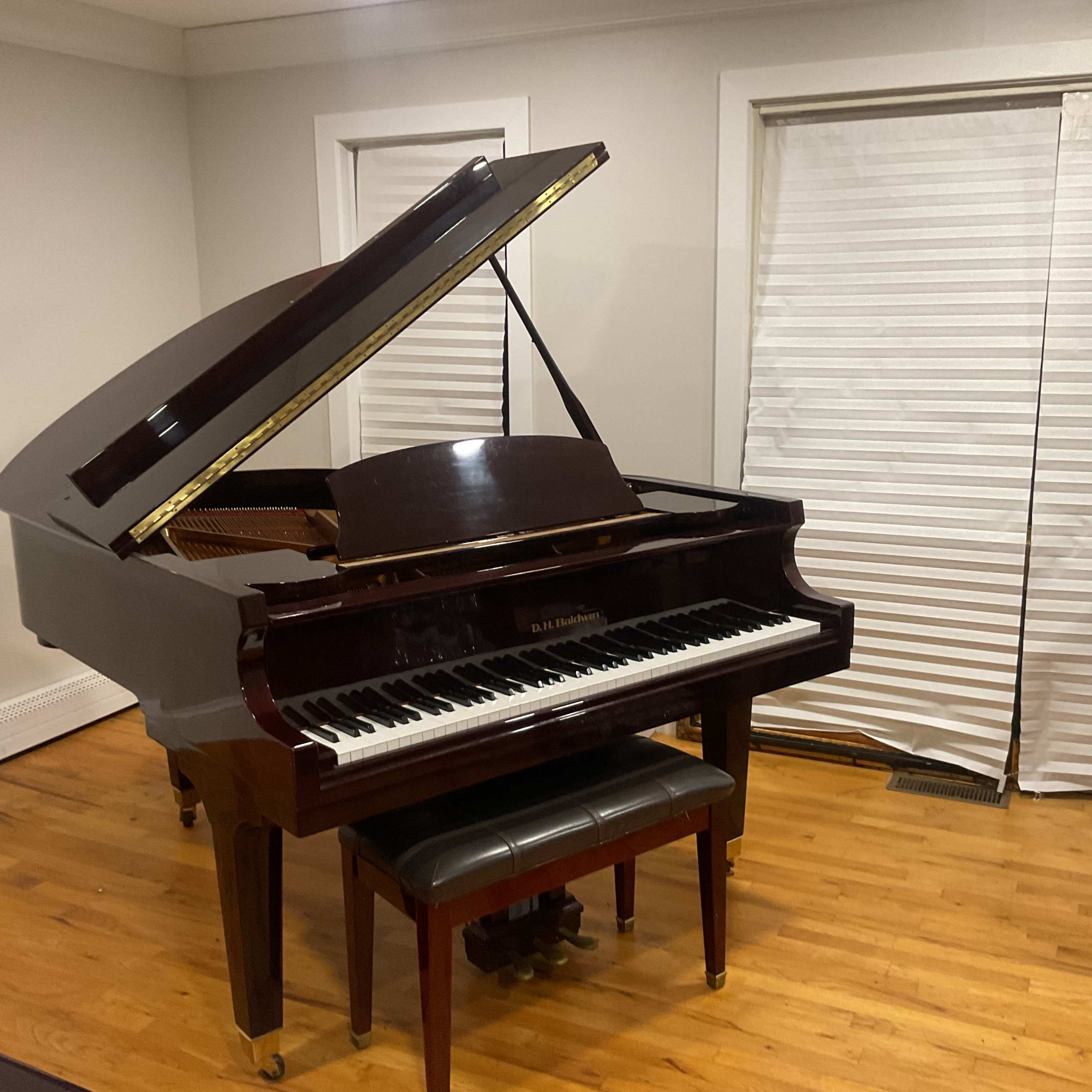 A grand piano with an open lid sits on polished wooden flooring beside closed window blinds.