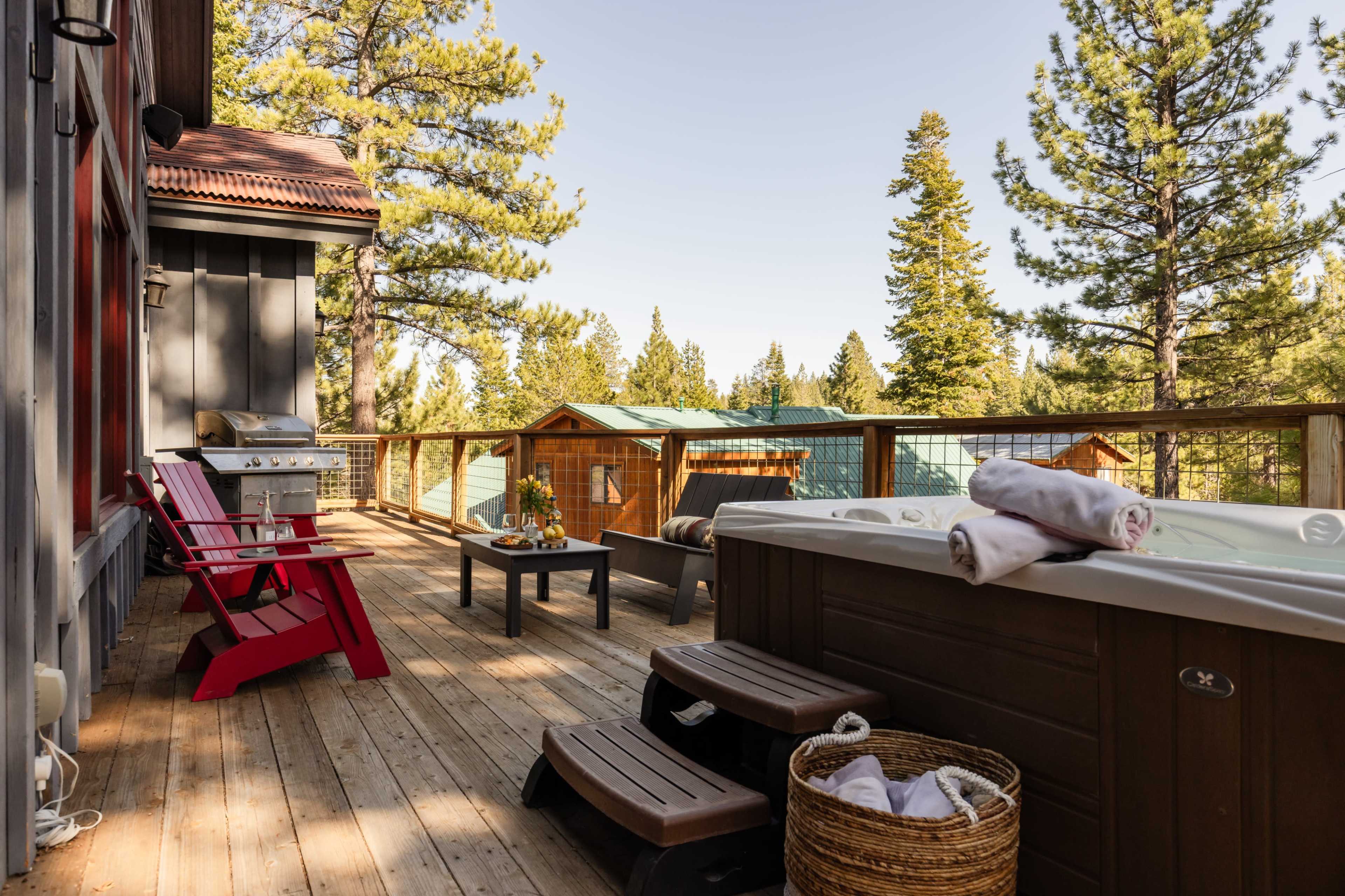 The image shows a deck with a hot tub, outdoor seating, and a grill, surrounded by tall pine trees and a view of nearby cabins.