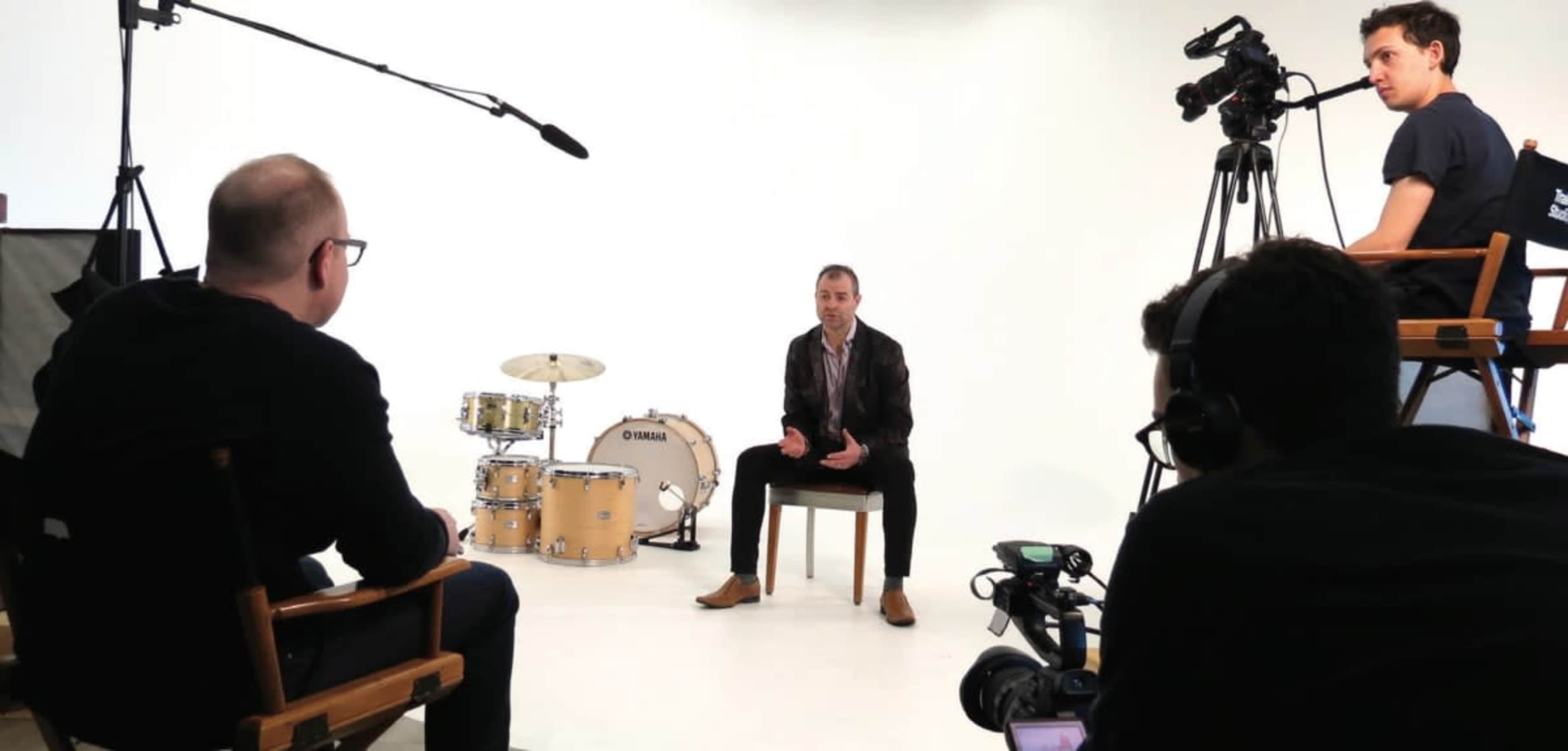 A man sits on a chair in front of a plain background while being interviewed, with cameras and lighting equipment set up around him.