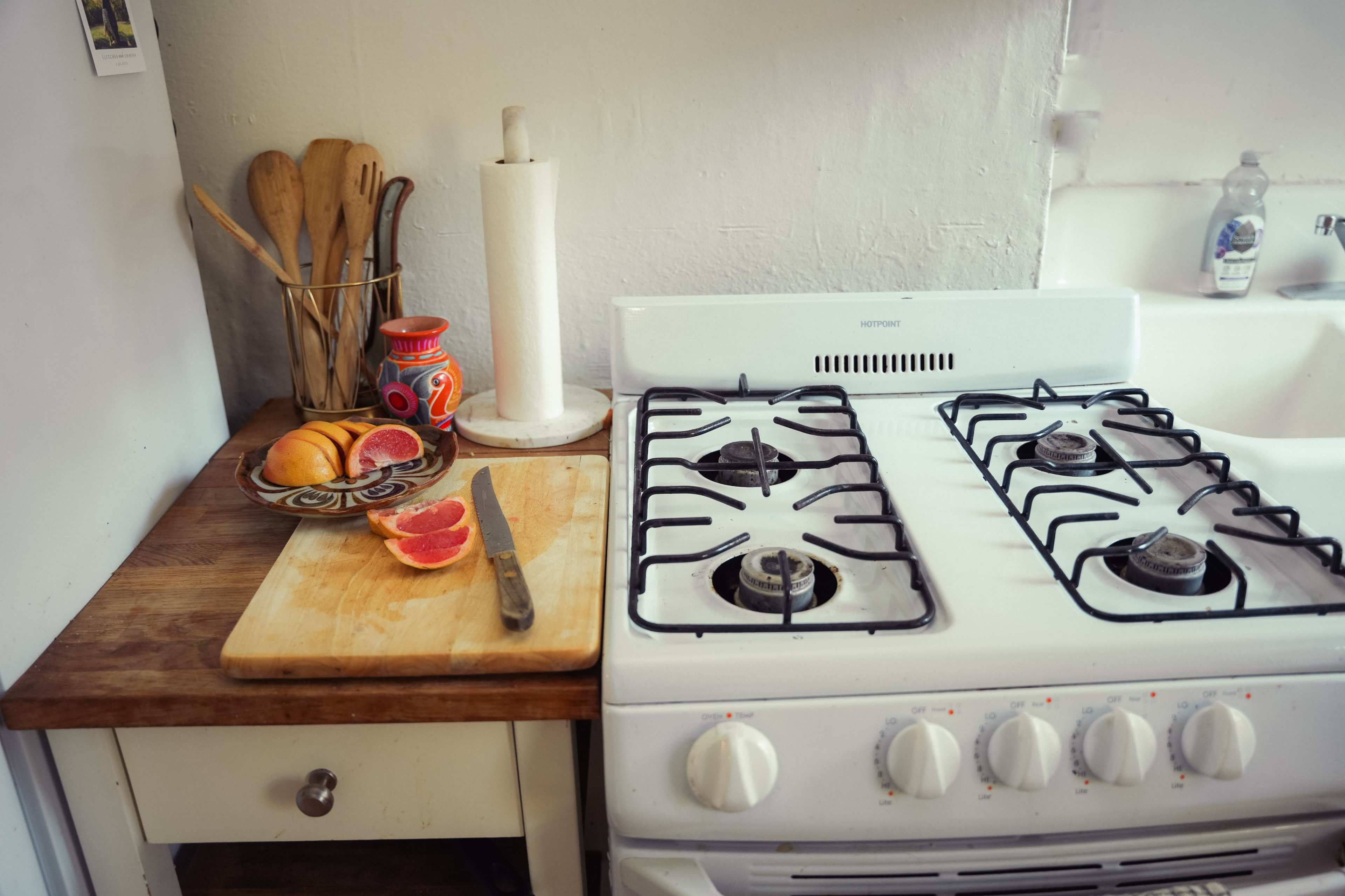 A wooden countertop features a cutting board with sliced grapefruit, a knife, a decorative bowl of fruit, a paper towel holder, and a white gas stove.