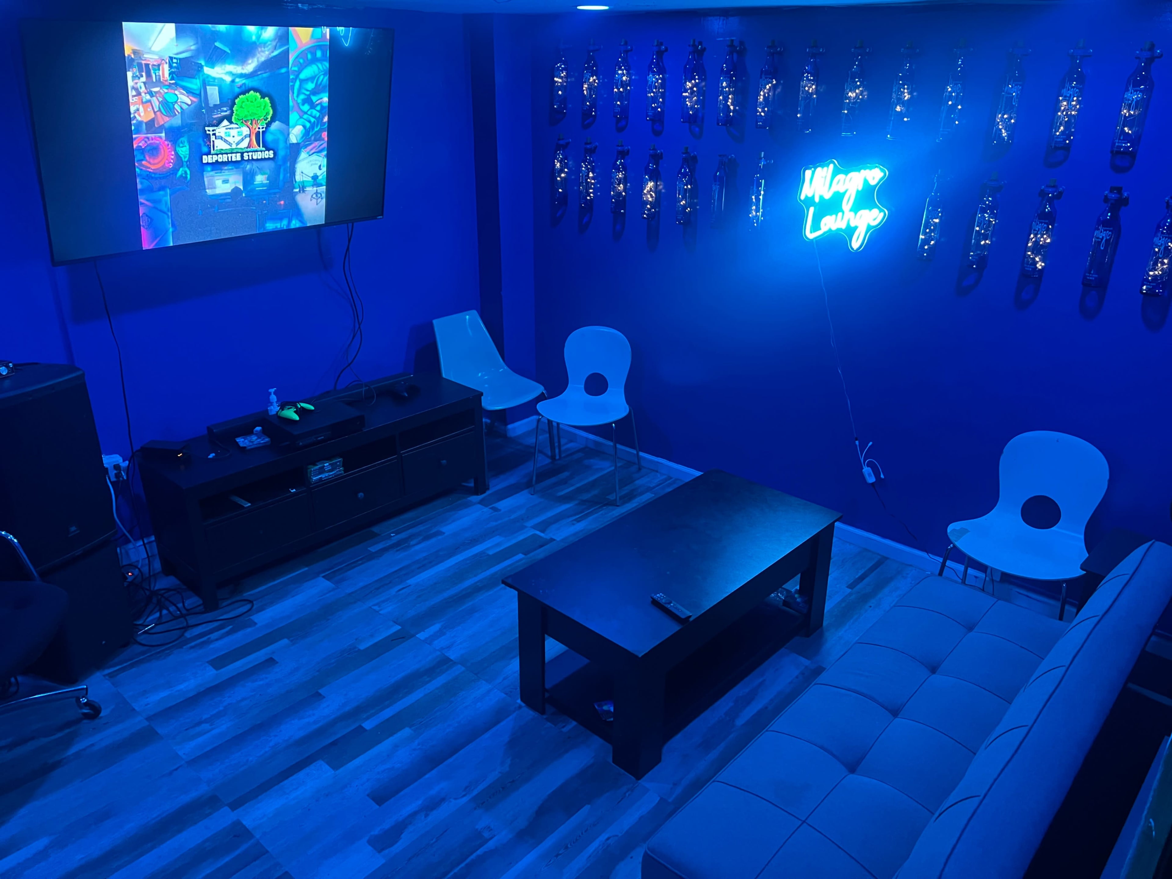 The room features a blue wall with neon signage, a large television displaying a game interface, and modern furniture including a sofa, chairs, and a coffee table.