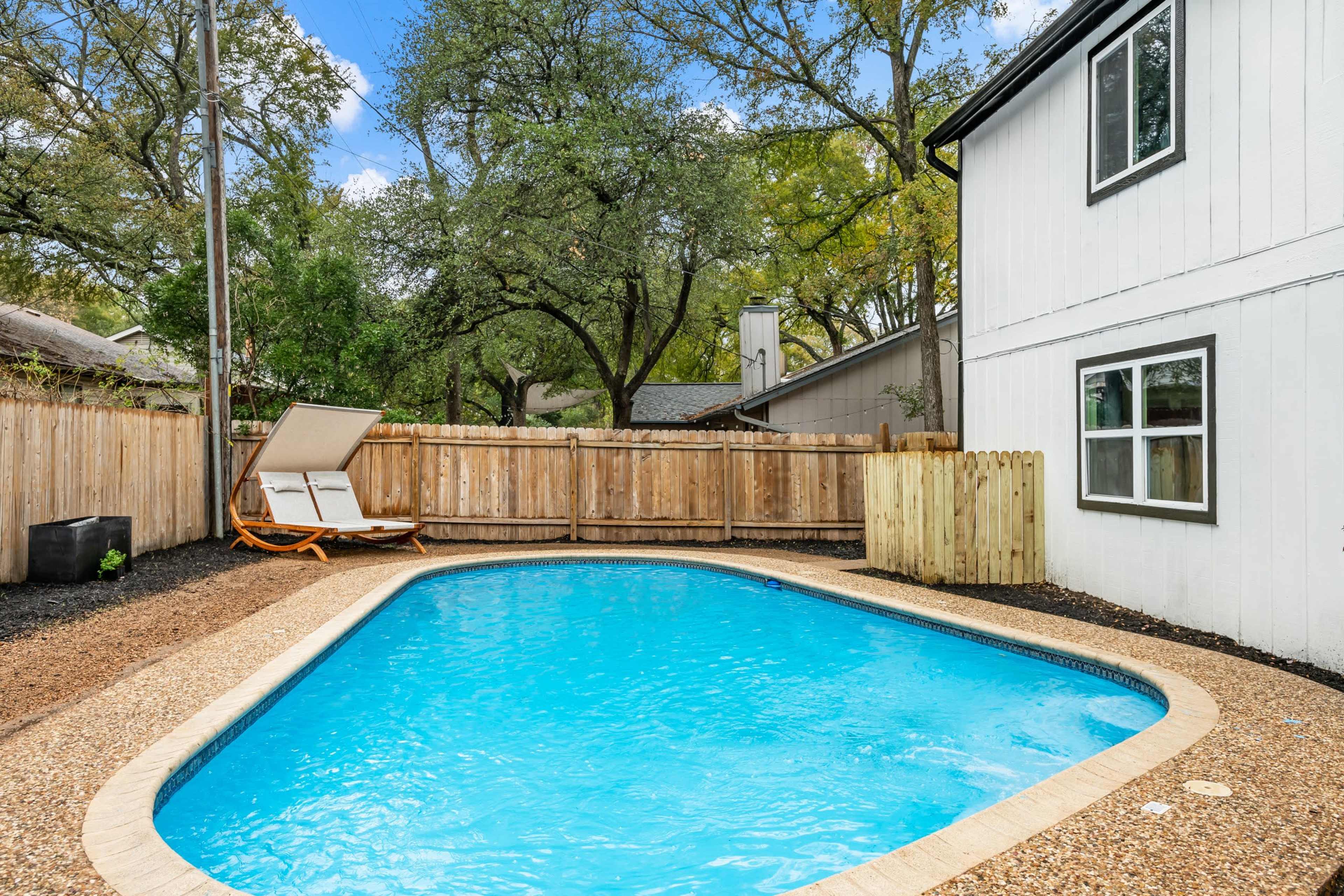 A rectangular swimming pool surrounded by a gravel area and a wooden fence, with a lounge chair placed nearby.
