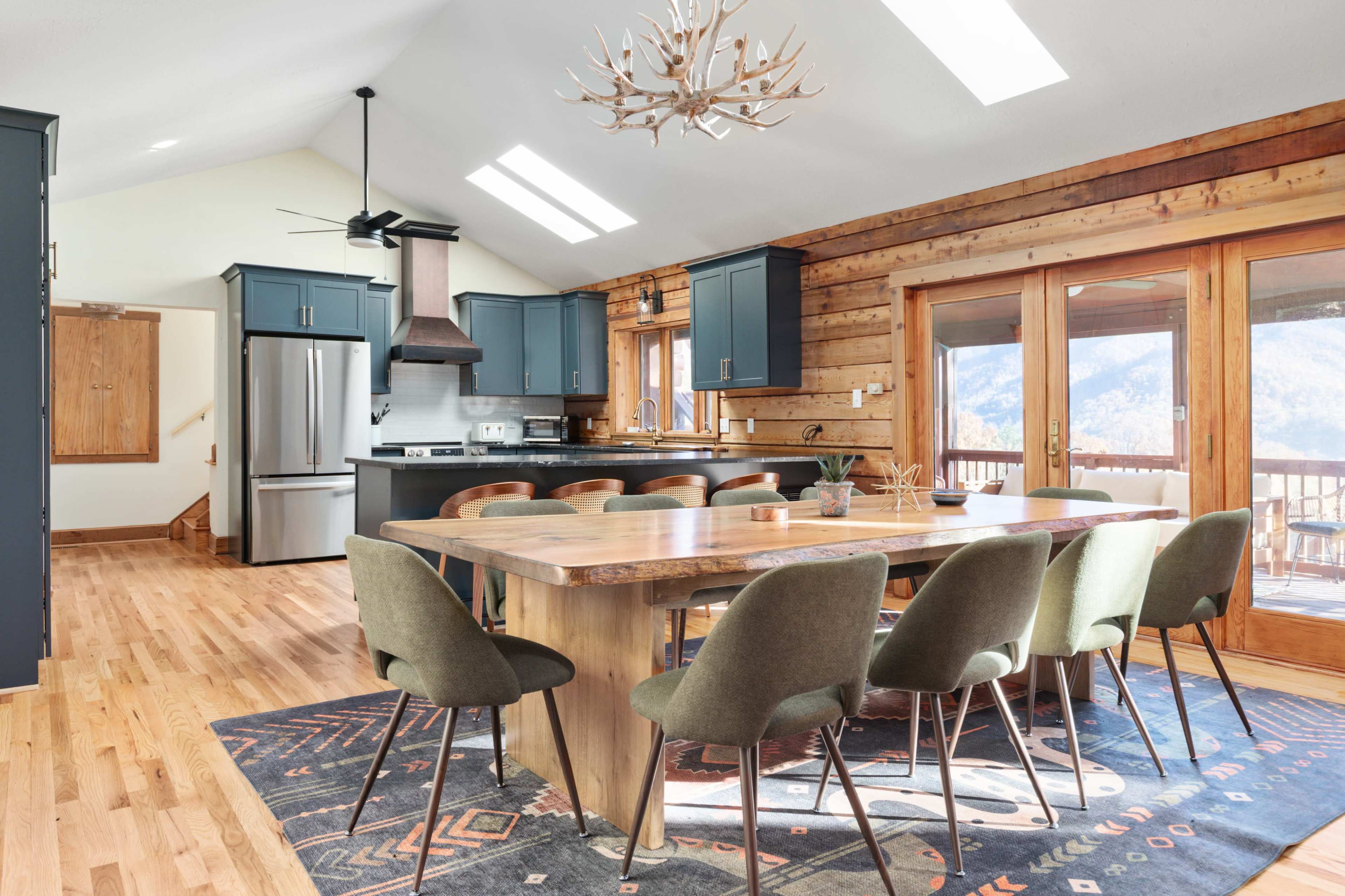 The image shows a modern kitchen and dining area with a wooden dining table, green upholstered chairs, and large windows that provide natural light and mountain views.