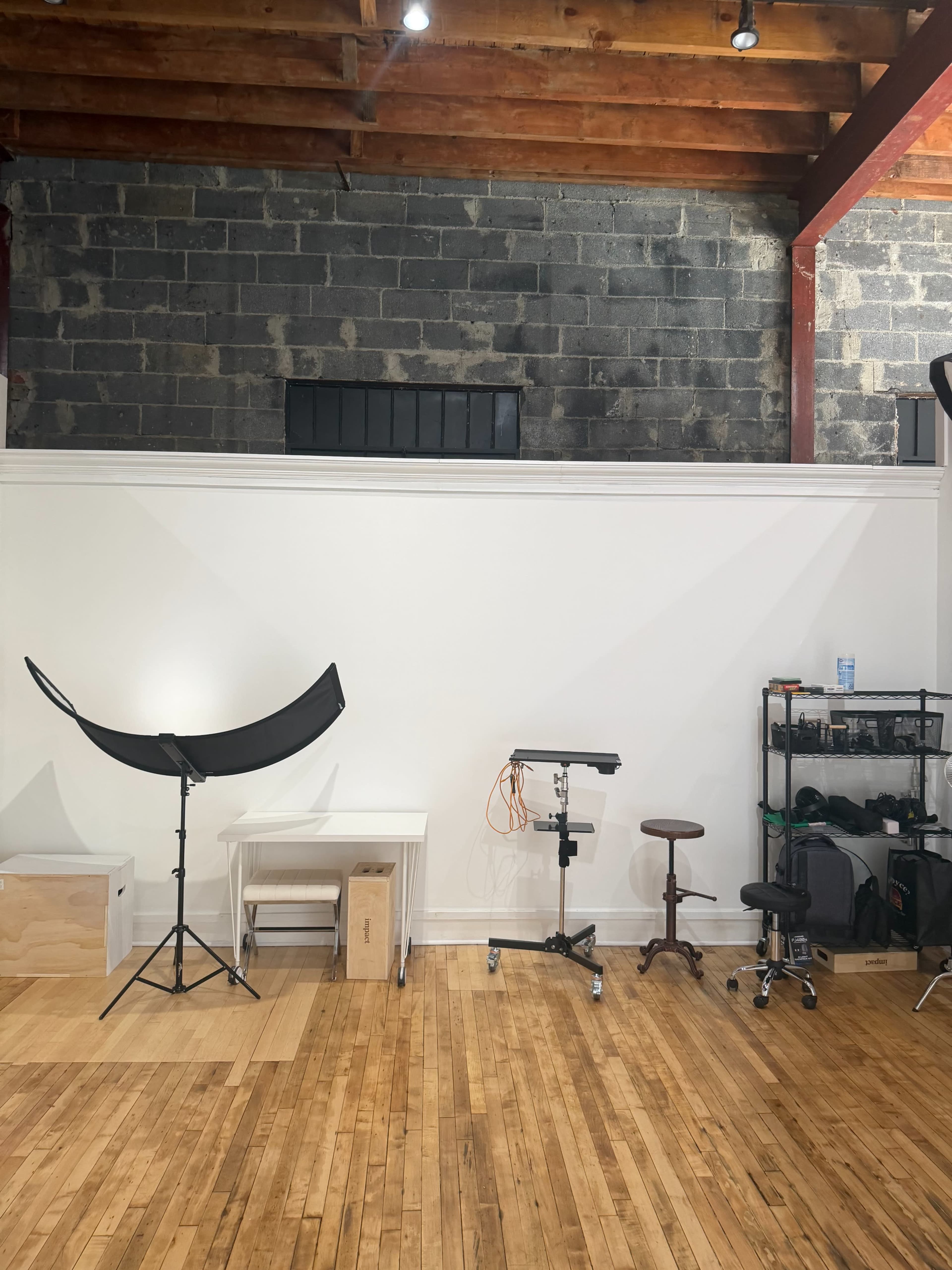 The image shows a spacious studio with hardwood floors, featuring a white wall, a photography backdrop setup, a desk, a rolling stool, and equipment on shelves.