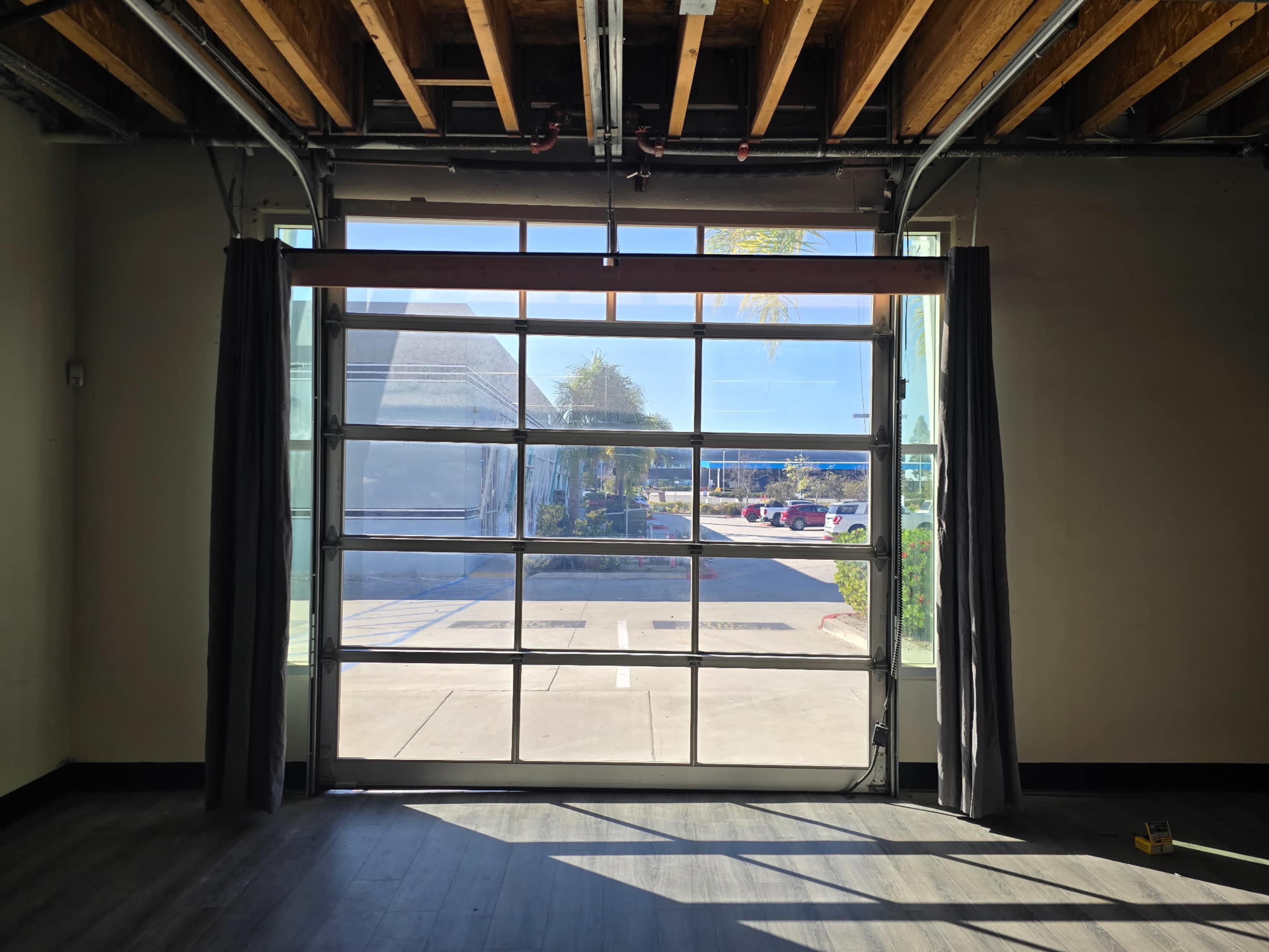 The image shows an open glass garage door with large panes, leading to a view of a parking lot and trees outside.
