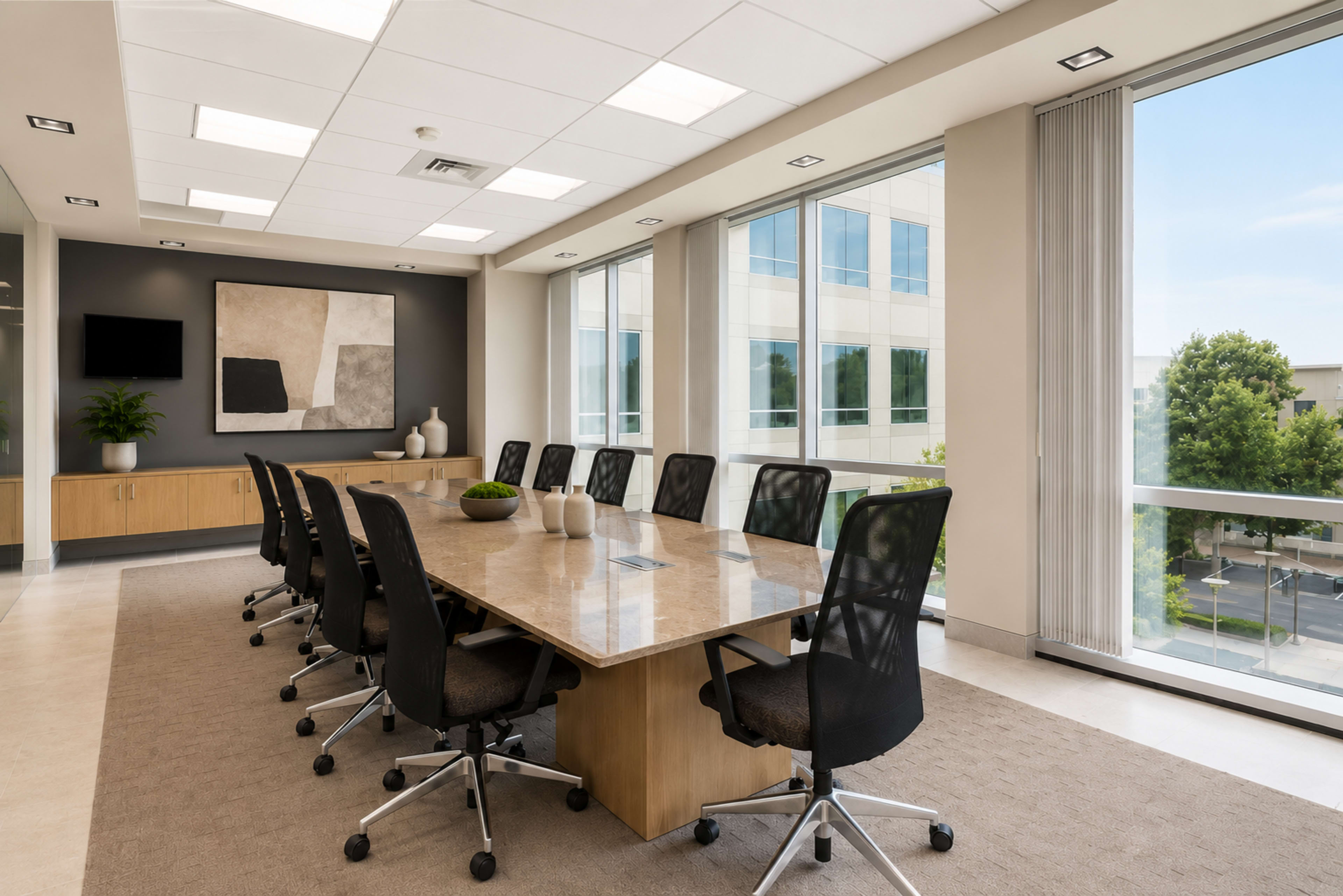 A spacious conference room features a long beige table surrounded by black rolling chairs, large windows providing natural light, and modern decor including a wall painting and plants.