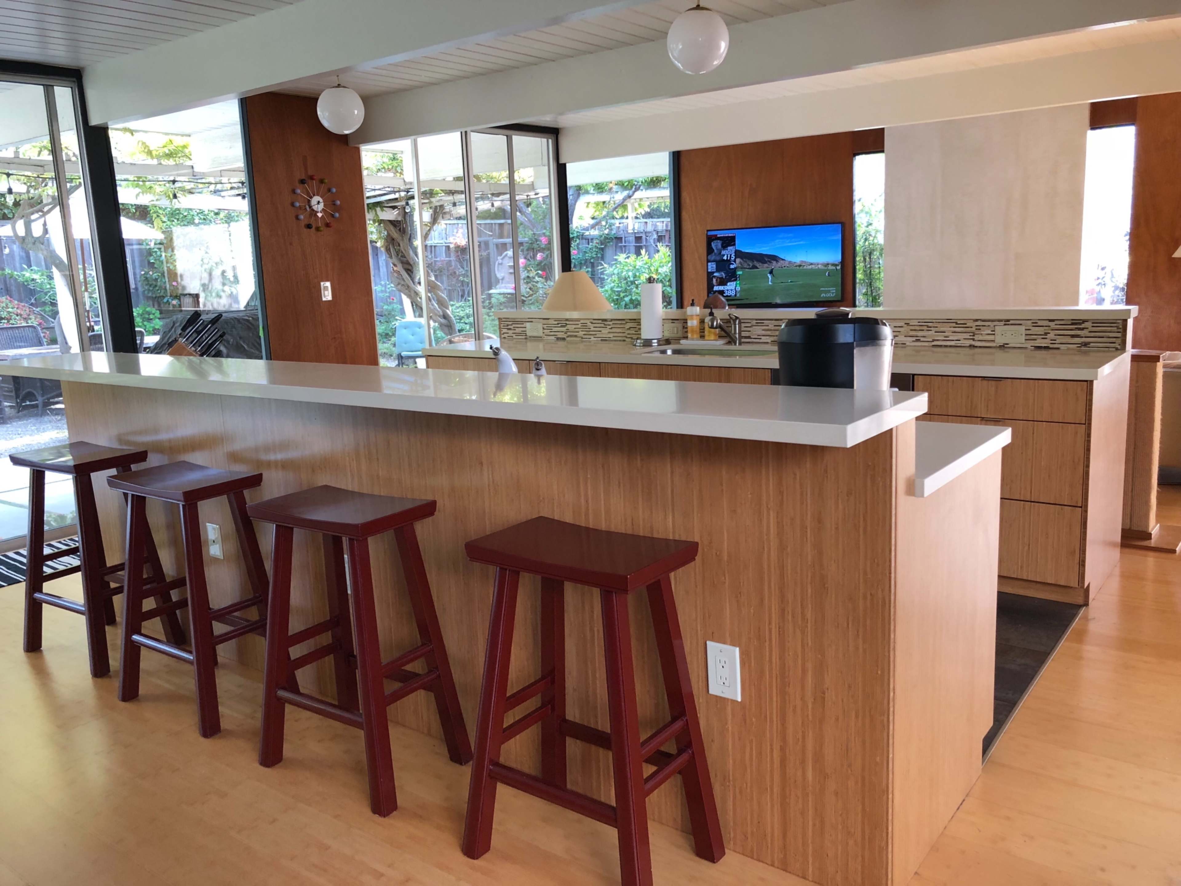 A modern kitchen with a bar counter and four wooden stools, featuring a television and windows that overlook a garden area.