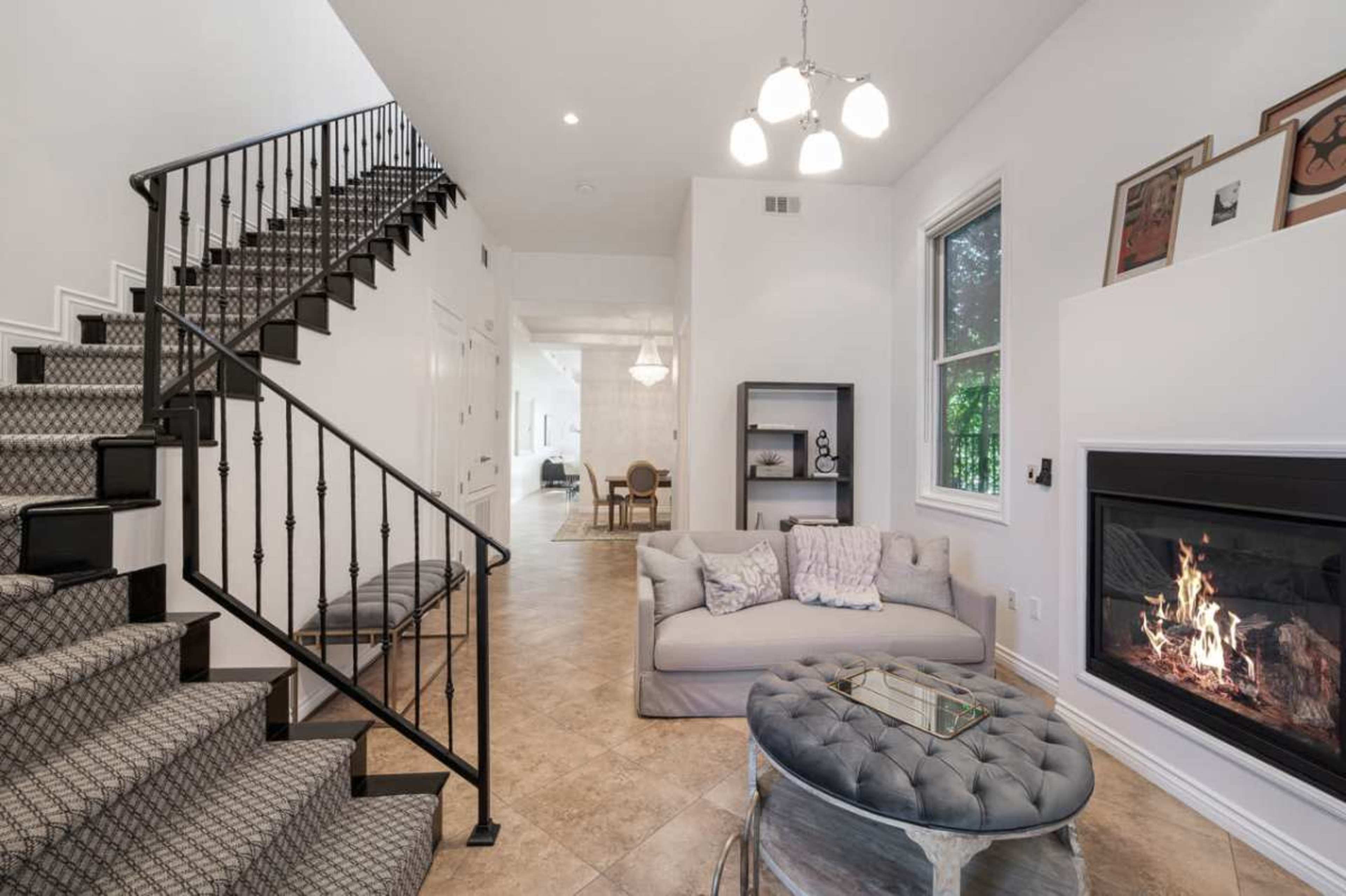 The image shows a modern living room with a staircase on the left, a circular ottoman in the center, and a fireplace on the wall to the right.
