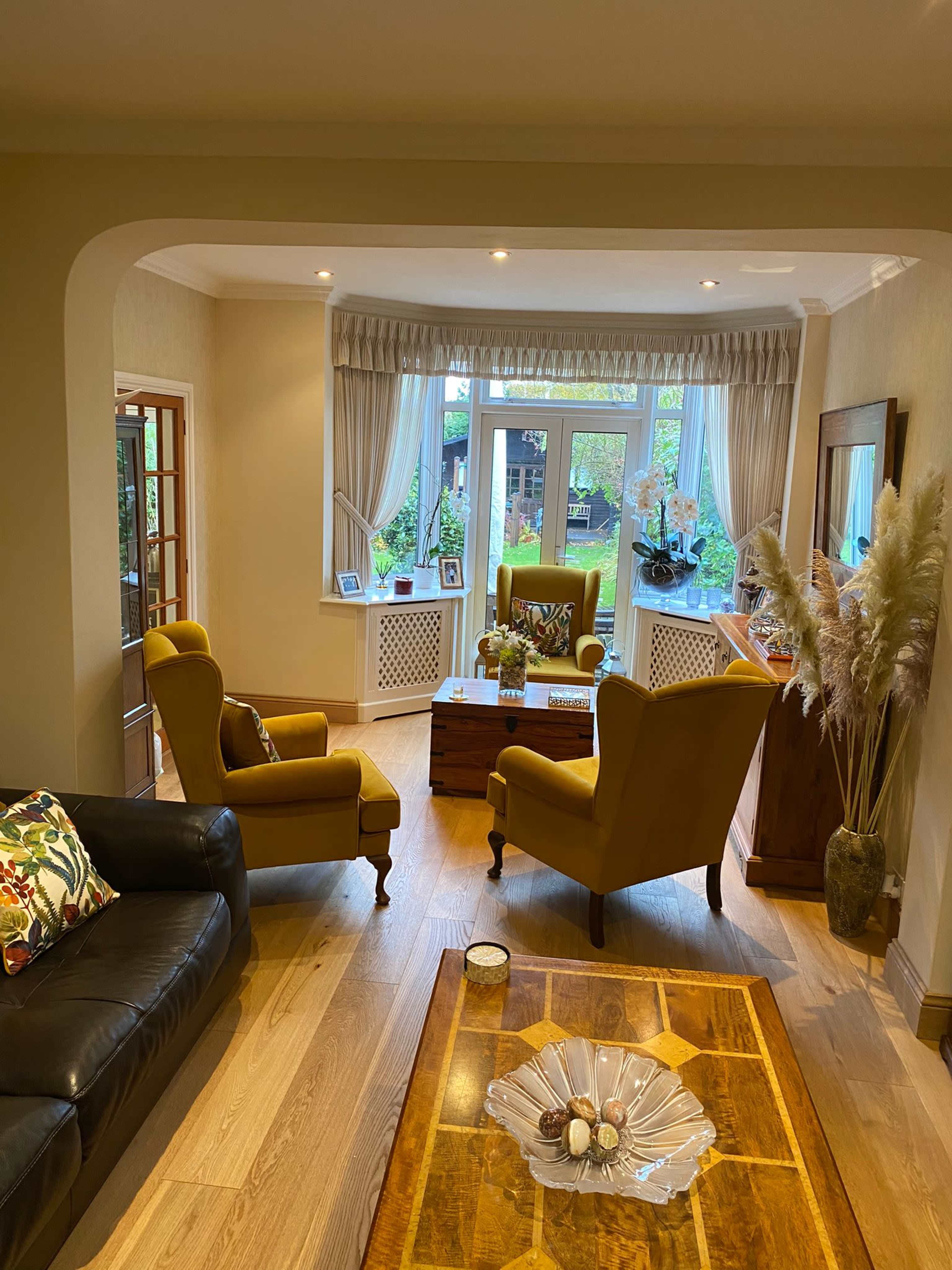 A living room with two yellow armchairs facing a wooden coffee table, a dark sofa, and large windows with a view of greenery outside.