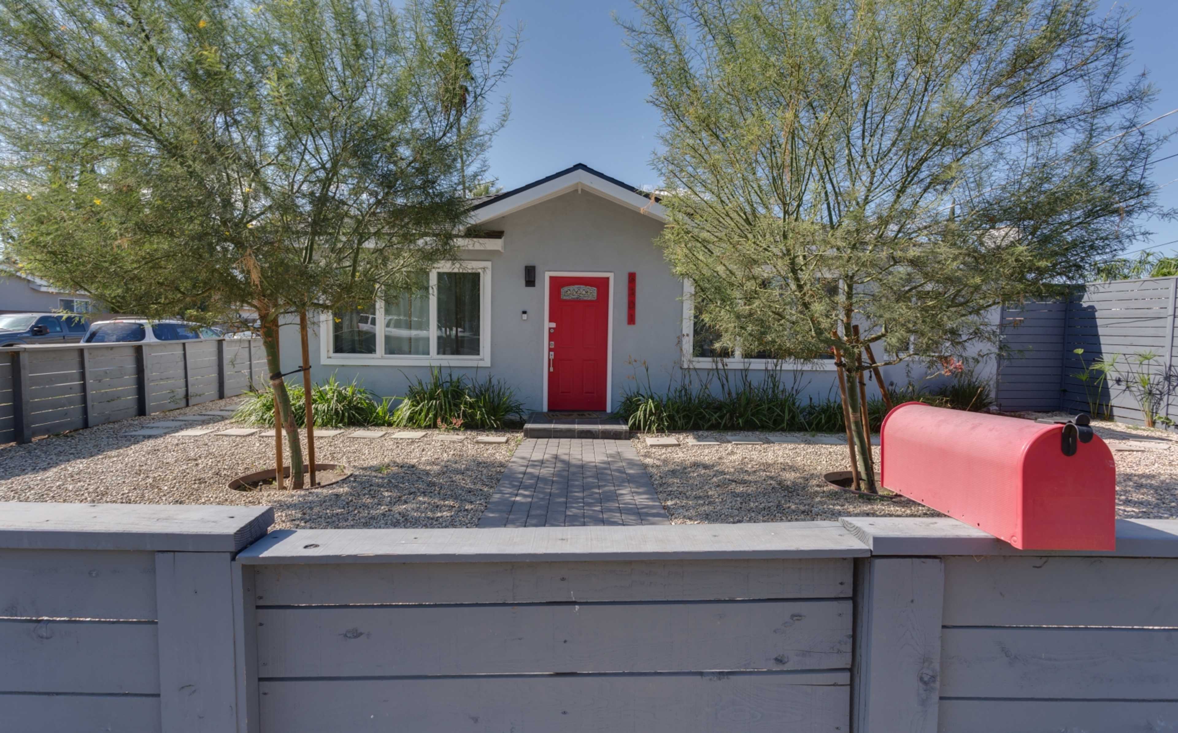 A gray house with a red front door is framed by two trees and a red mailbox, set within a gravel yard enclosed by a low wooden fence.