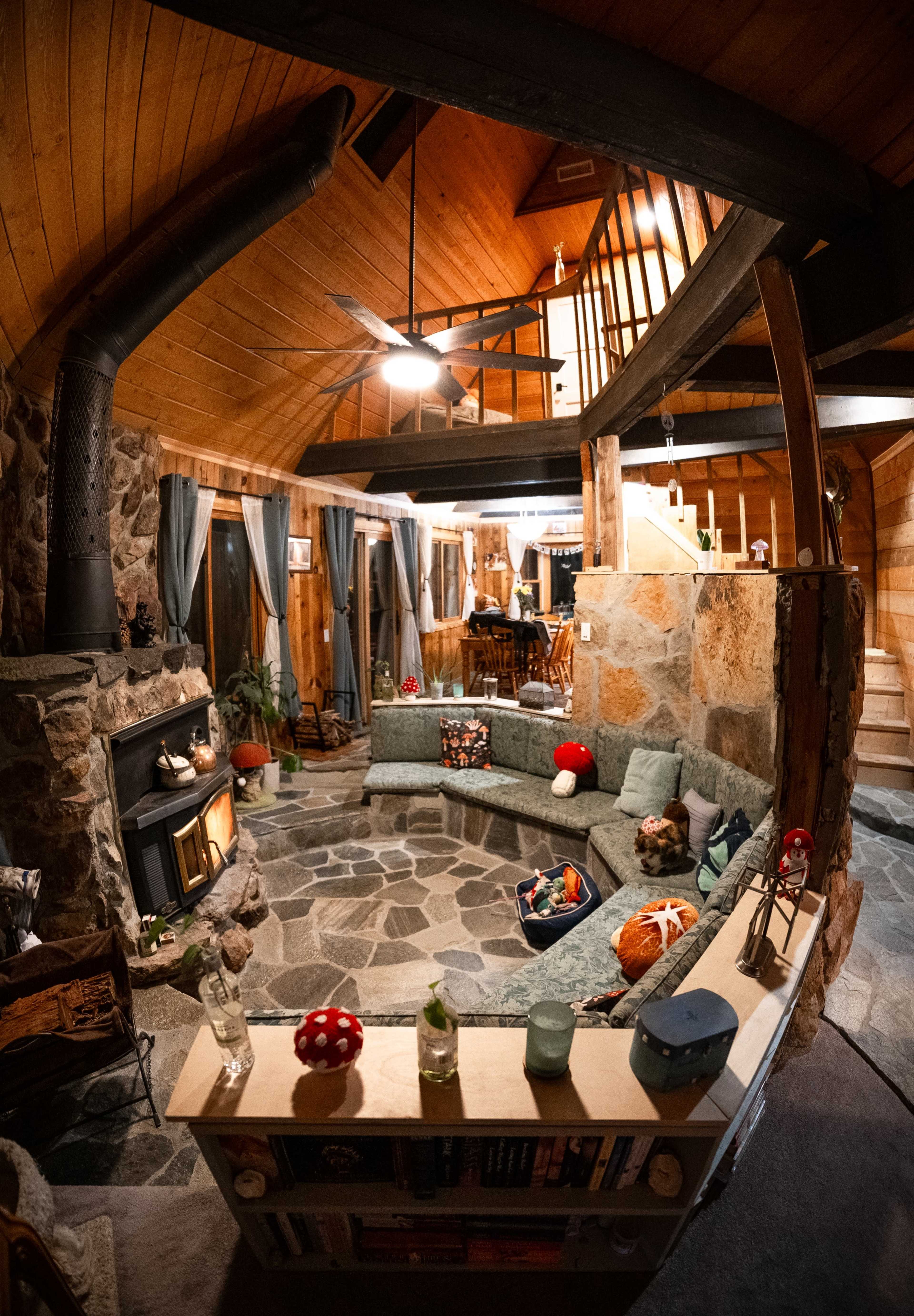 The interior of a rustic cabin features a circular seating area with a stone fireplace and wooden accents, surrounded by large windows and a staircase leading to an upper level.